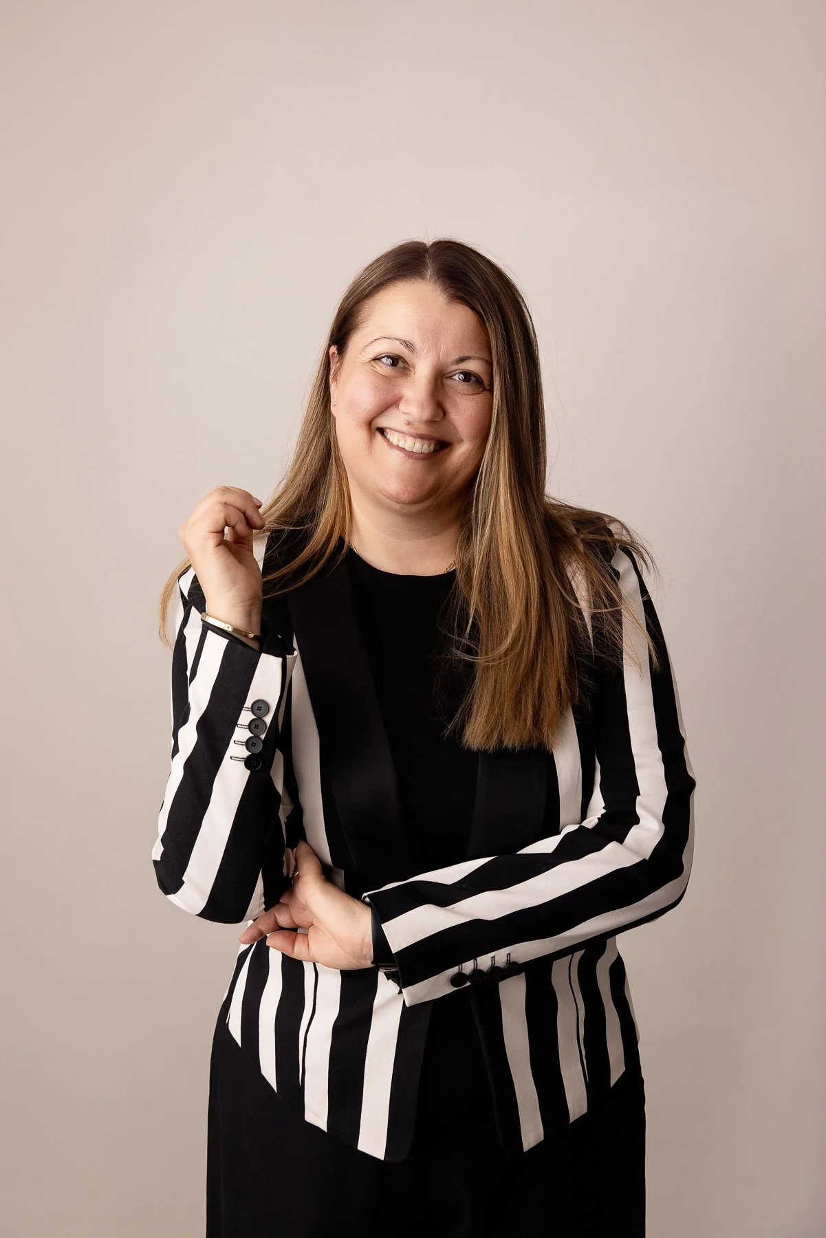 A woman with blonde hair wearing a black & white jacket, standing up smiling at the camera in a cozy, well-lit room.