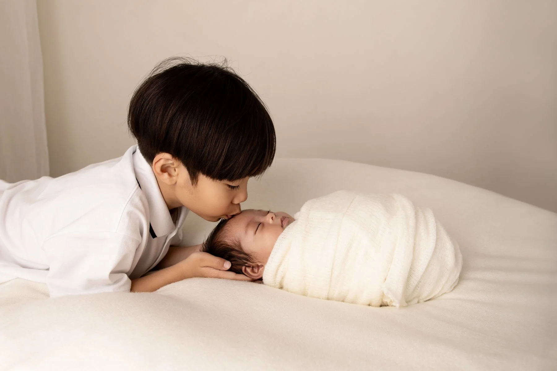 Young boy leaning over to kiss a swaddled baby on a white blanket.