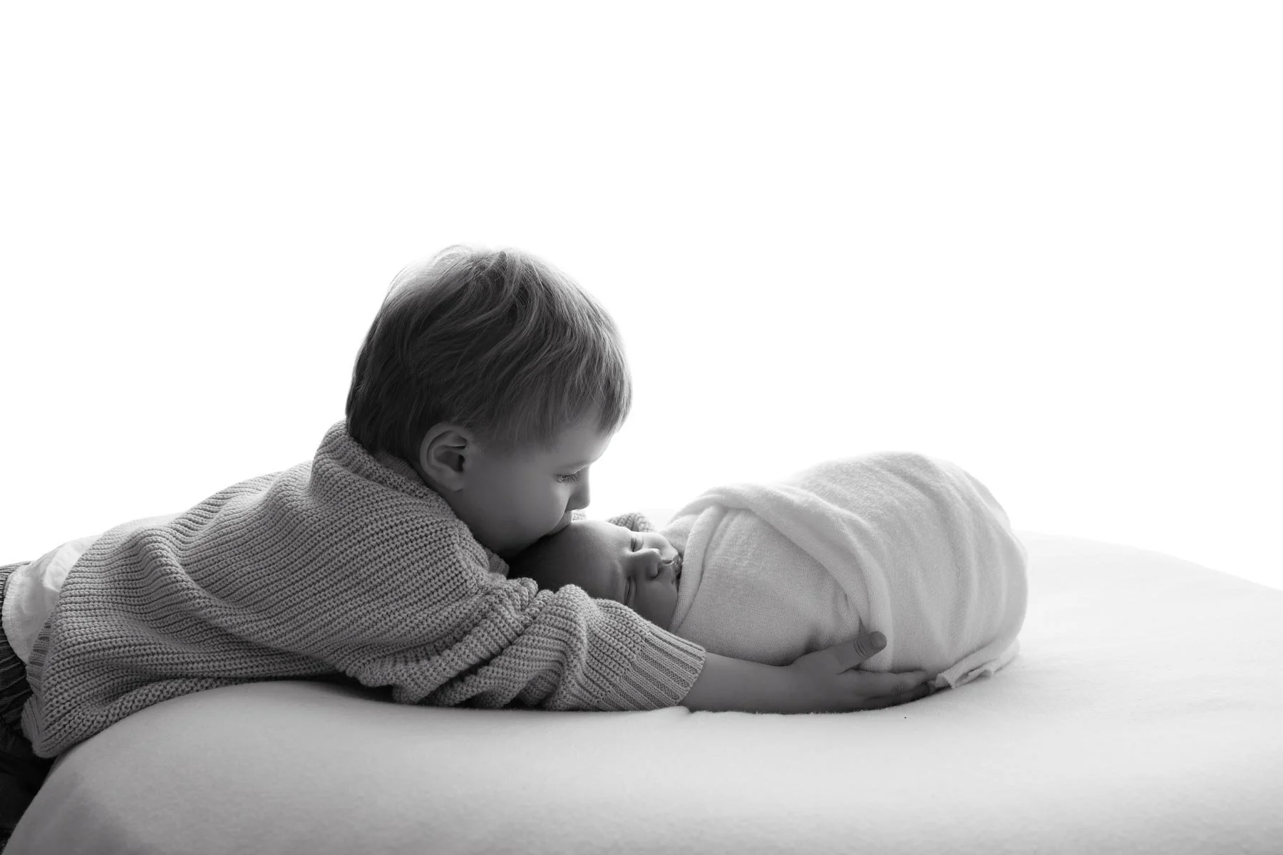 Young boy leaning over to kiss a swaddled baby on a white blanket. Black and white portrait