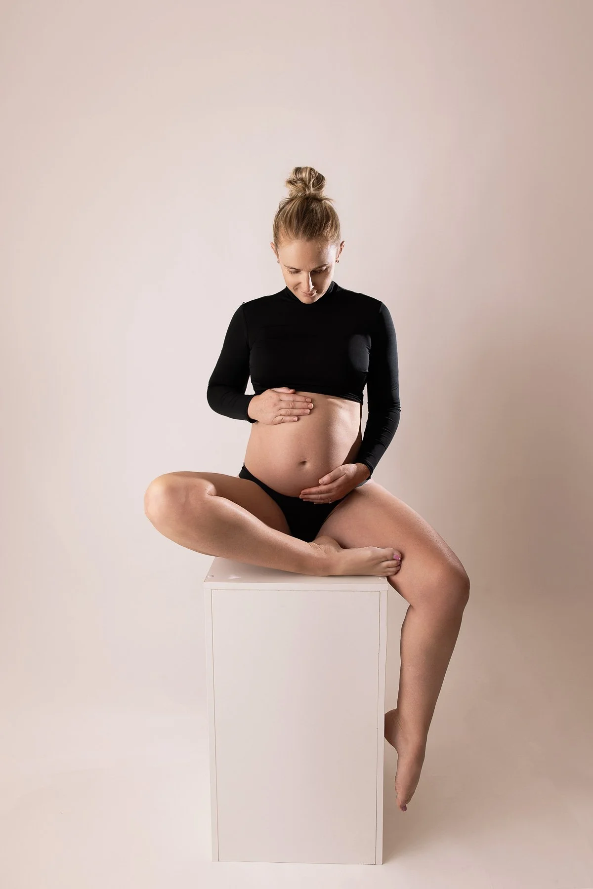 Maternity photo woman wearing a black crop top sitting on a white posing block, blond hair