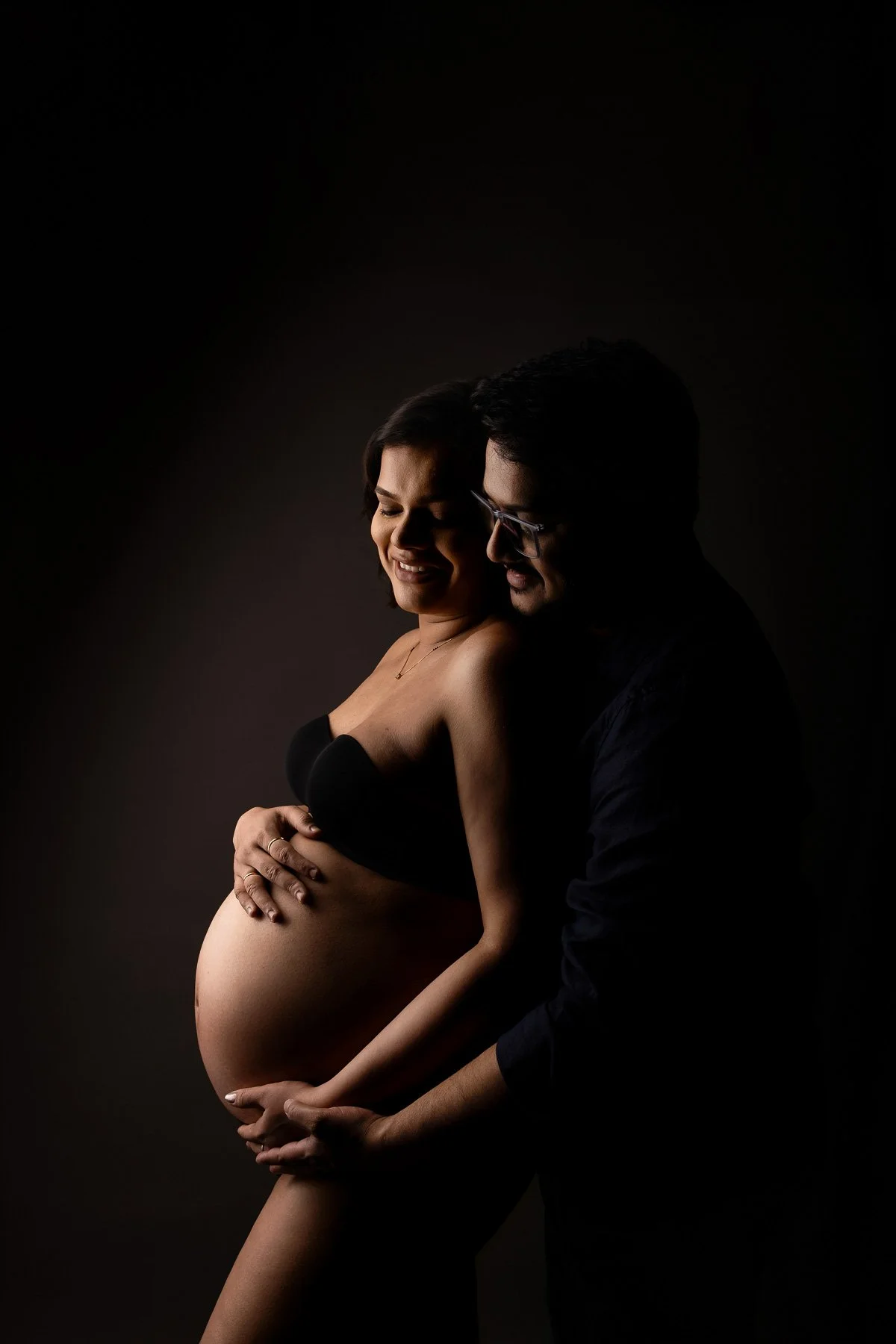 Pregnant woman in black maternity wear, w ith her husband holding her belly against a dark background. maternity photographer North Shore Sydney.