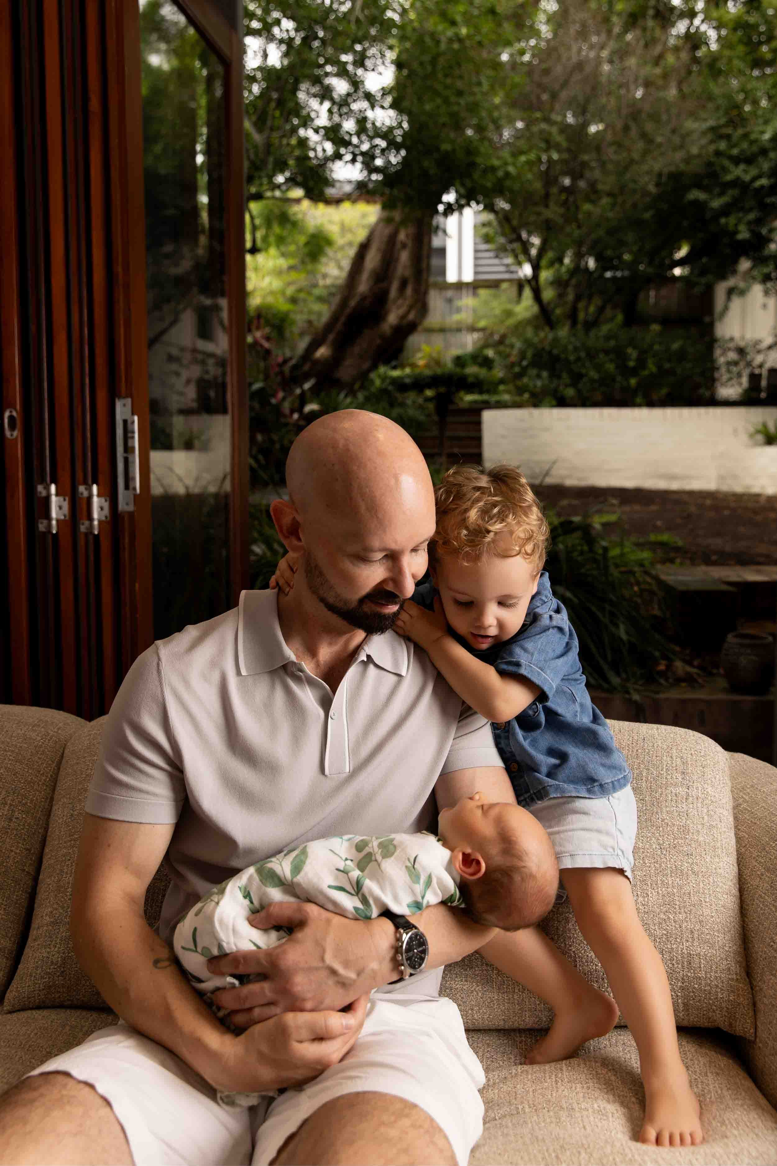 Man sitting on a couch holding a baby, with a young child hugging him, in a cozy living room with a garden view.