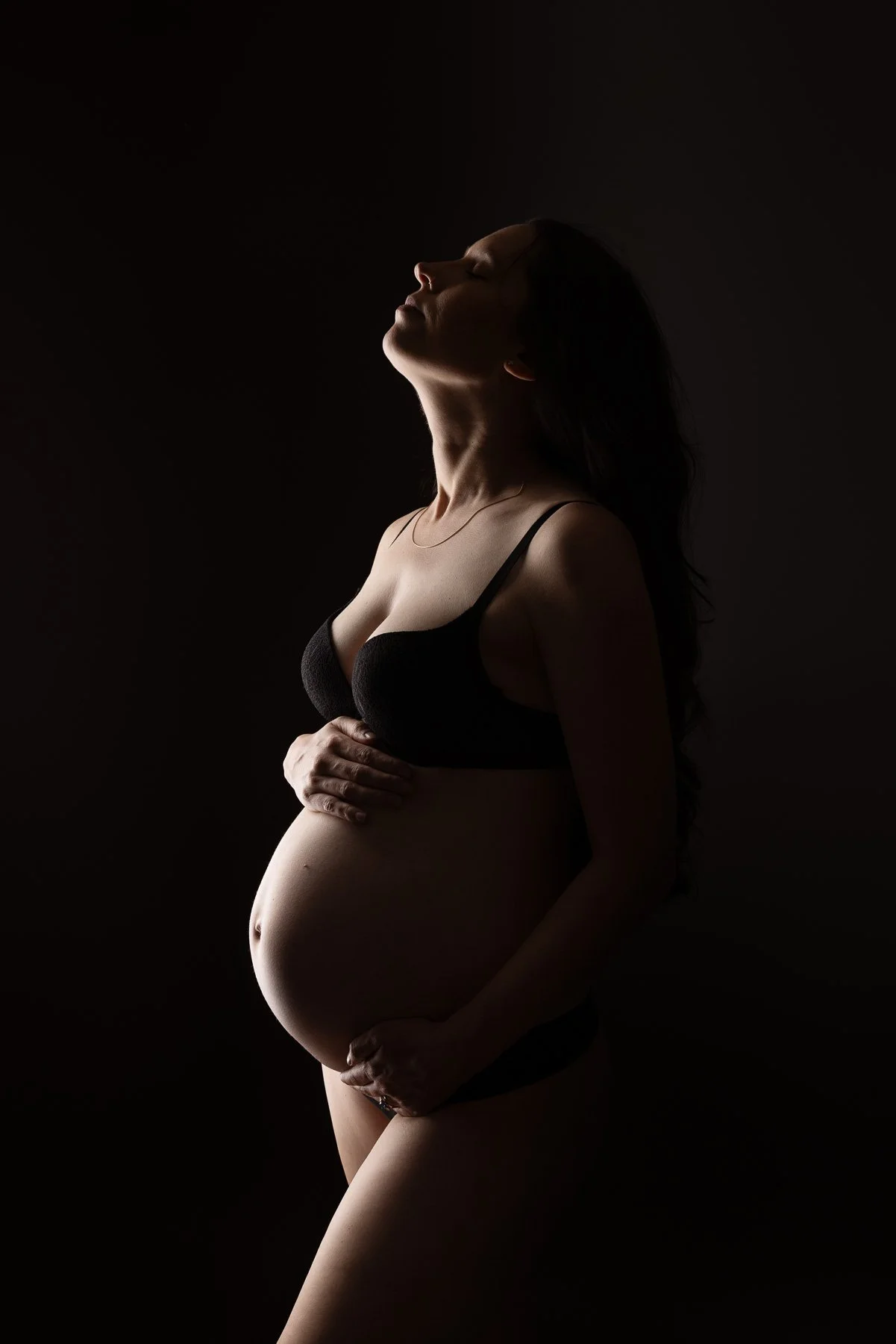 Pregnant woman in black maternity wear, holding her belly against a dark background. maternity photographer North Shore Sydney.
