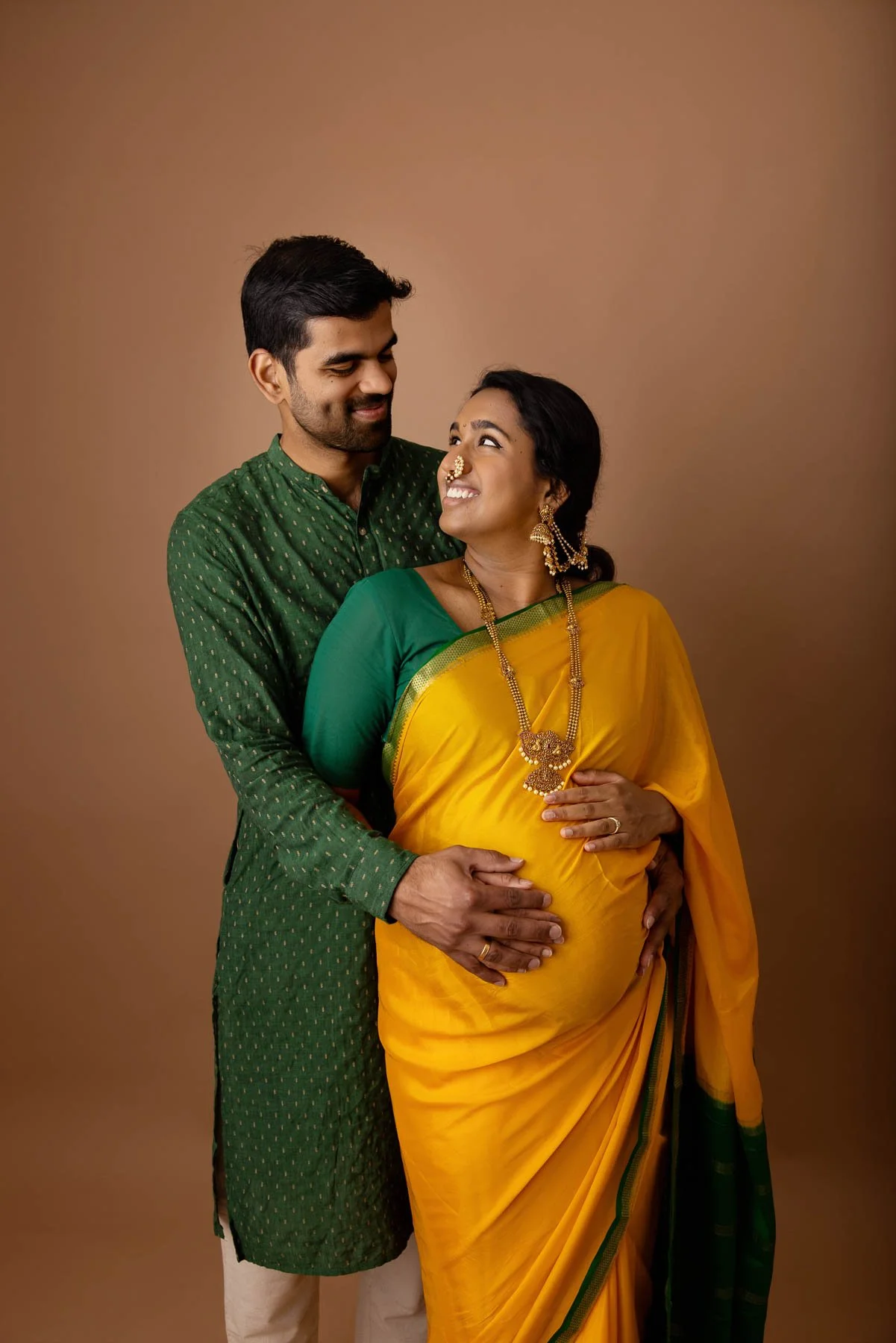 Couple smiling at each other. Pregnant woman in yellow & green saree, wearing golden jewelries, holding her belly, against a warm neutral background.
