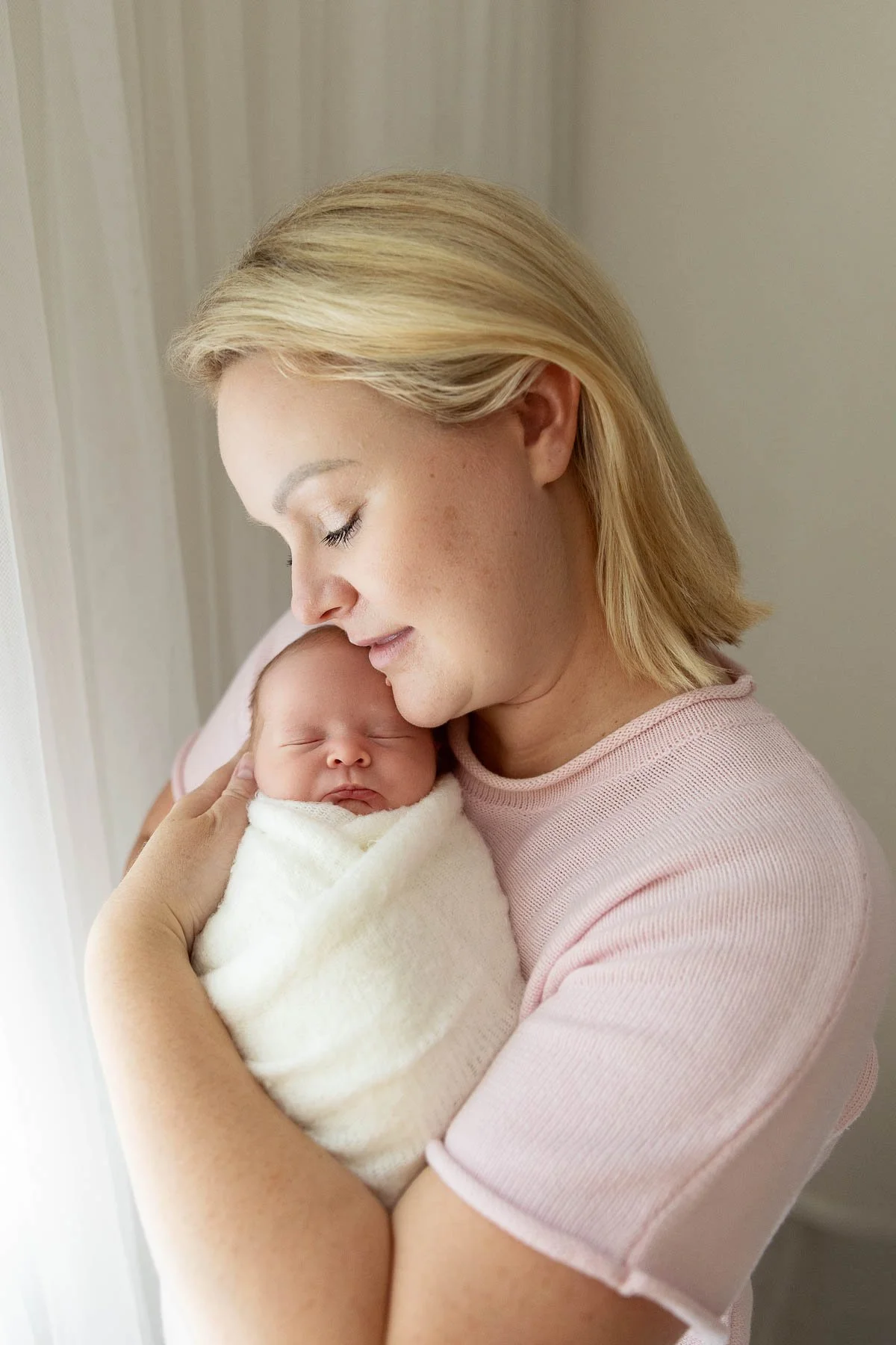 Mother holding close her newborn baby wrapped in white. Smiling.