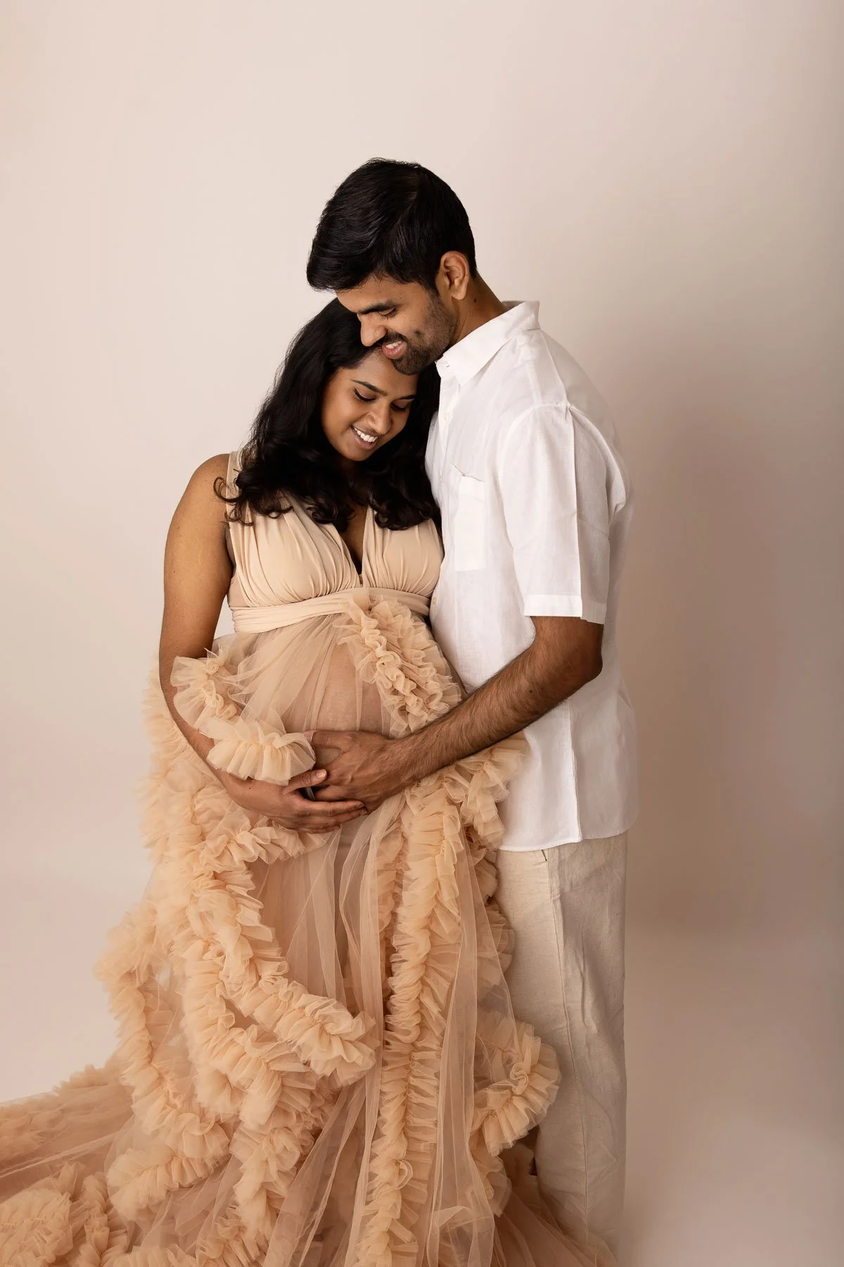 Pregnant couple in studio photo with white background, man touching woman's belly. The lady is wearing a big tulle dress.
