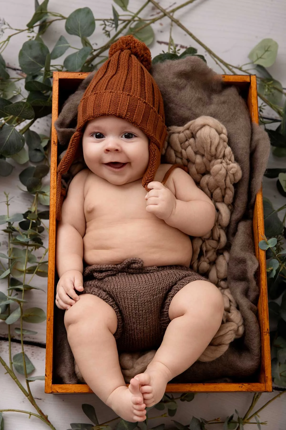Smiling baby wearing a knitted brown hat and shorts, lying in a wooden box with soft blankets and eucalyptus leaves around.