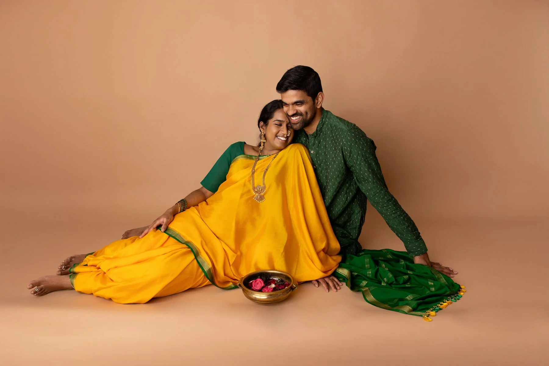 Pregnant woman in yellow & green saree, wearing golden jewelries, holding her belly, against a warm neutral background.