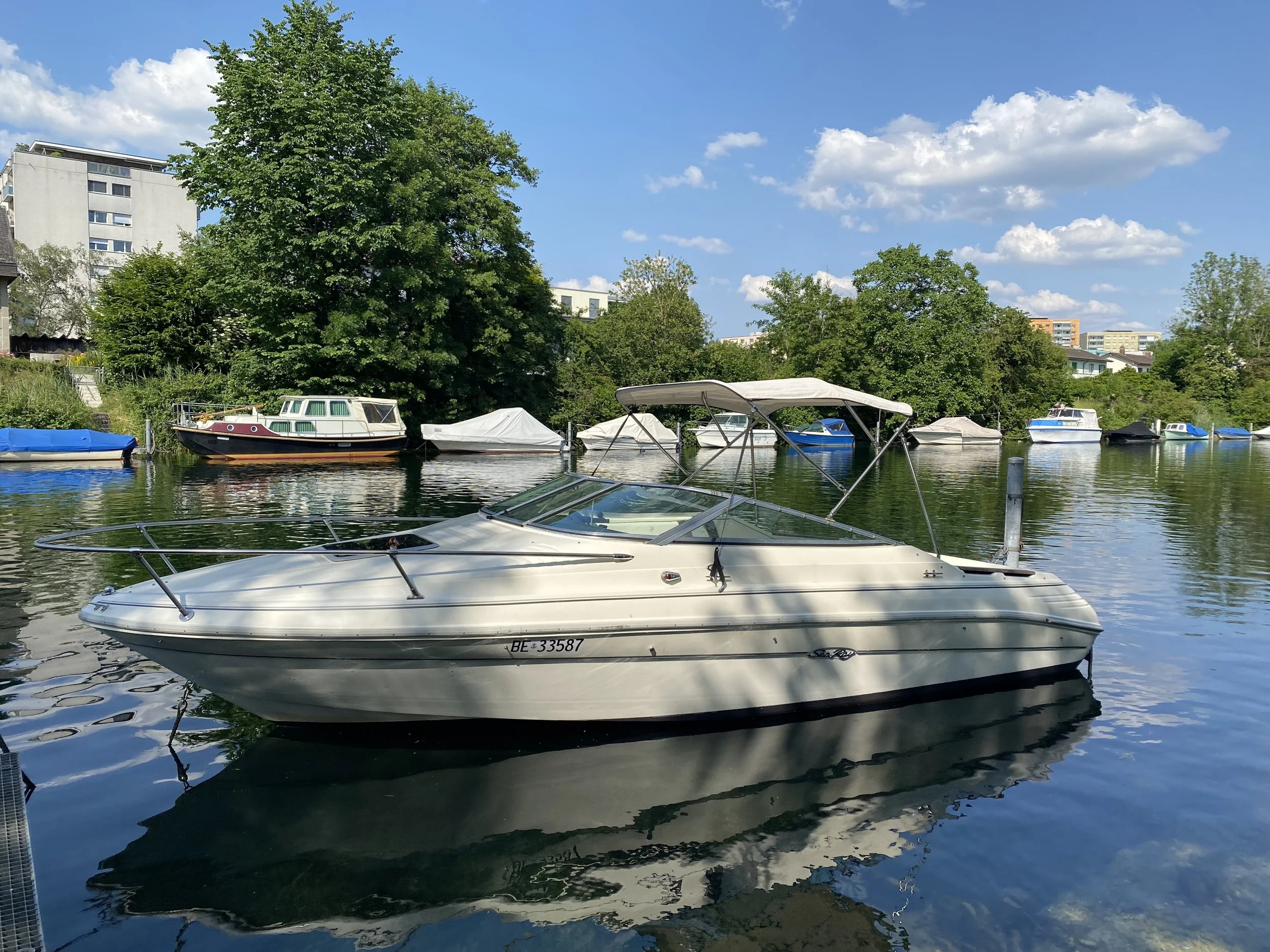 Weisses Mietboot an seinem Liegeplatz im Bielersee mit offenem, schattenspendenden Sonnenverdeck. Im Hintergrund sind verschiedene Boote an ihren Liegeplätzen zu sehen.
