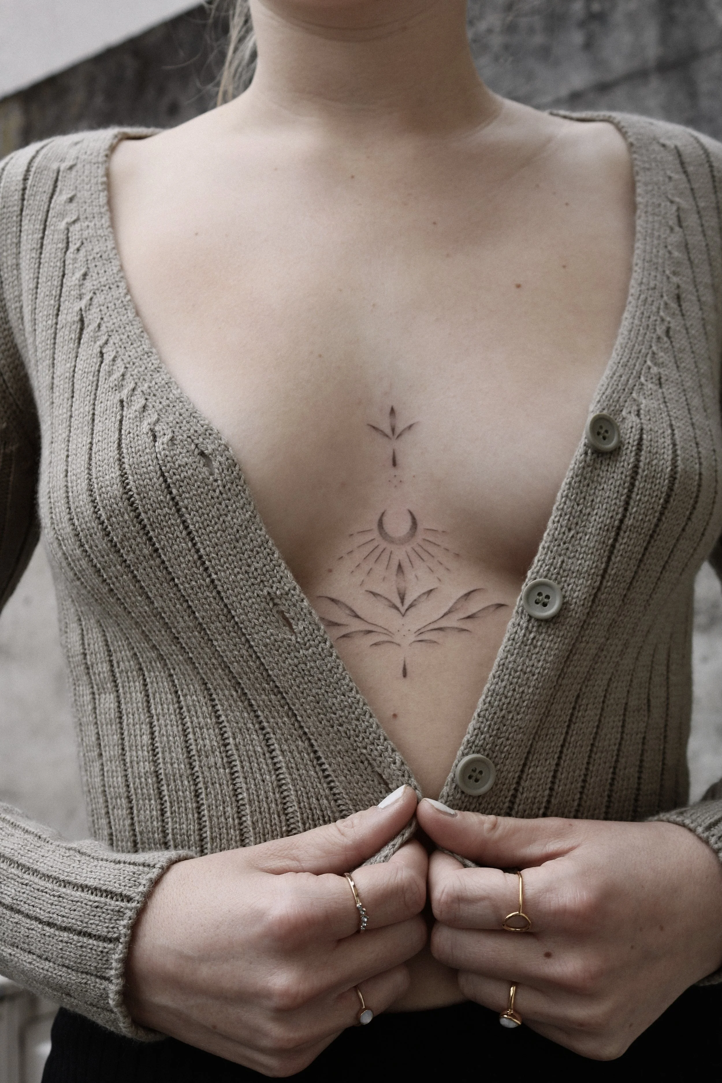 Oberkörper einer Person mit beigefarbener Strickjacke und geometrischem Tattoo auf der Brust.