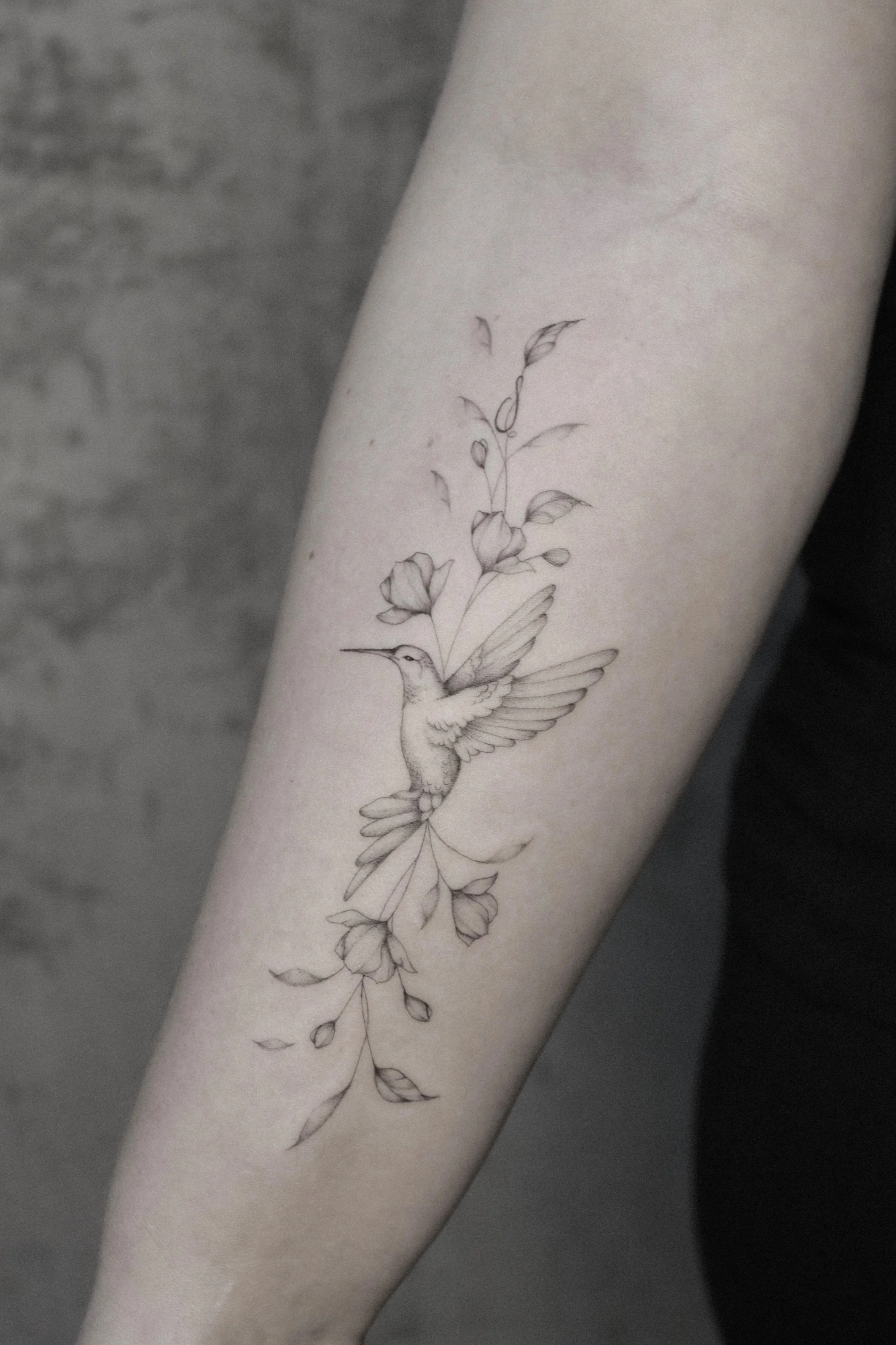 Tätowierung eines Kolibris mit Blumen auf dem Arm einer Person.