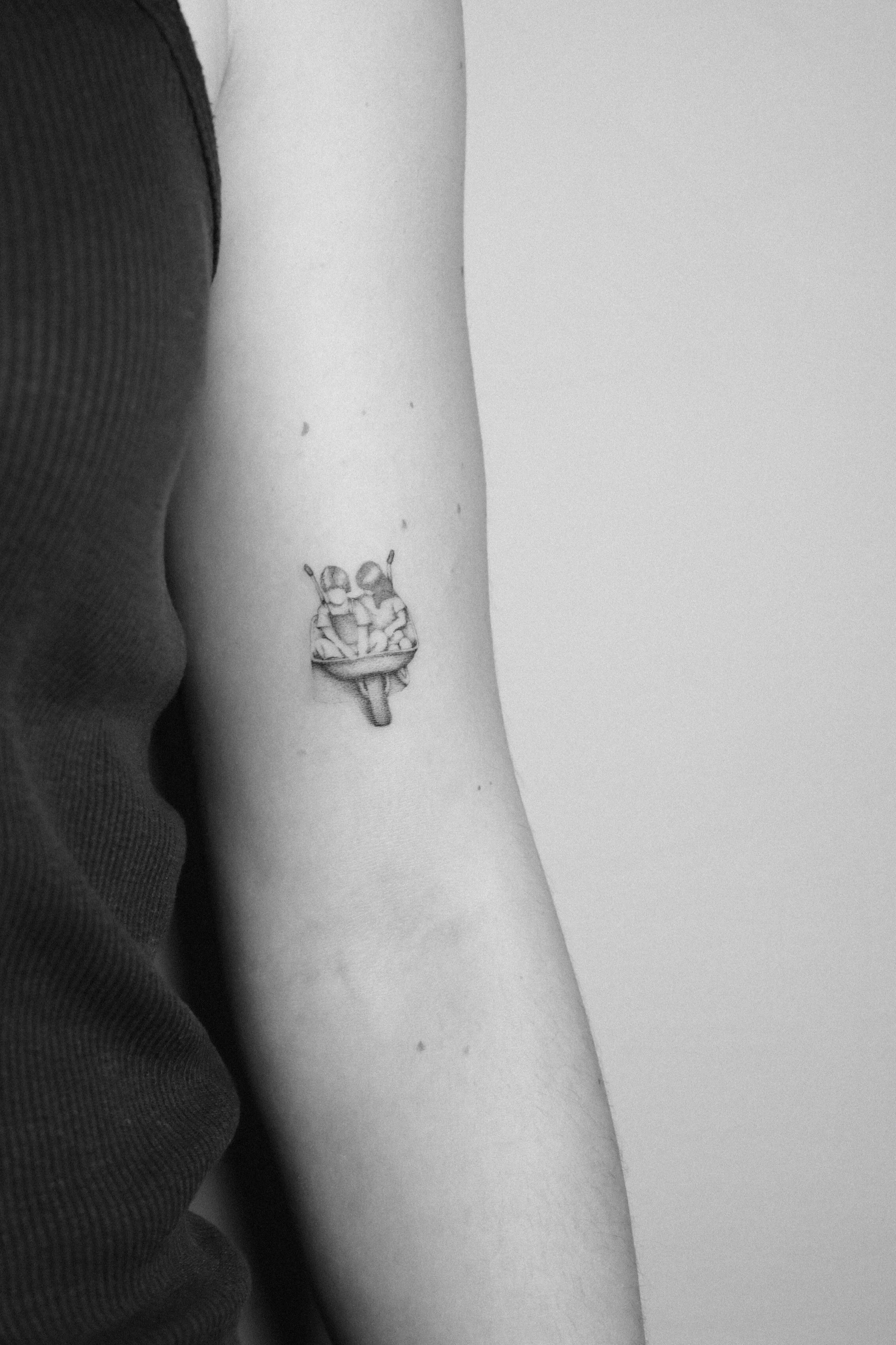 Schwarz-Weiß-Bild eines Oberarms mit einem kleinen, detaillierten Tattoo von zwei Menschen in einem Boot.