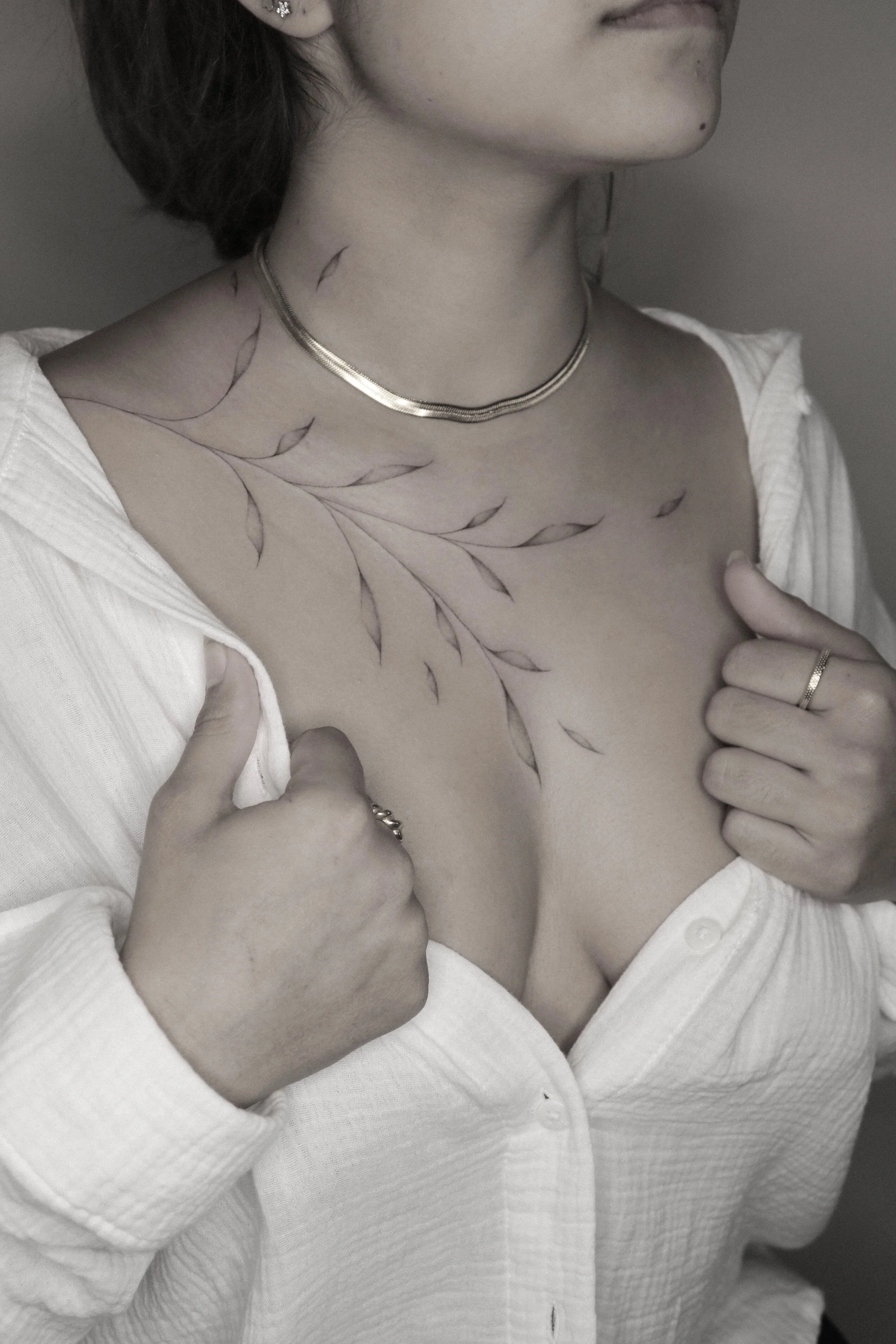 Frau mit Blätter-Tattoo am Dekolleté, goldene Halskette, weißes Hemd leicht geöffnet.