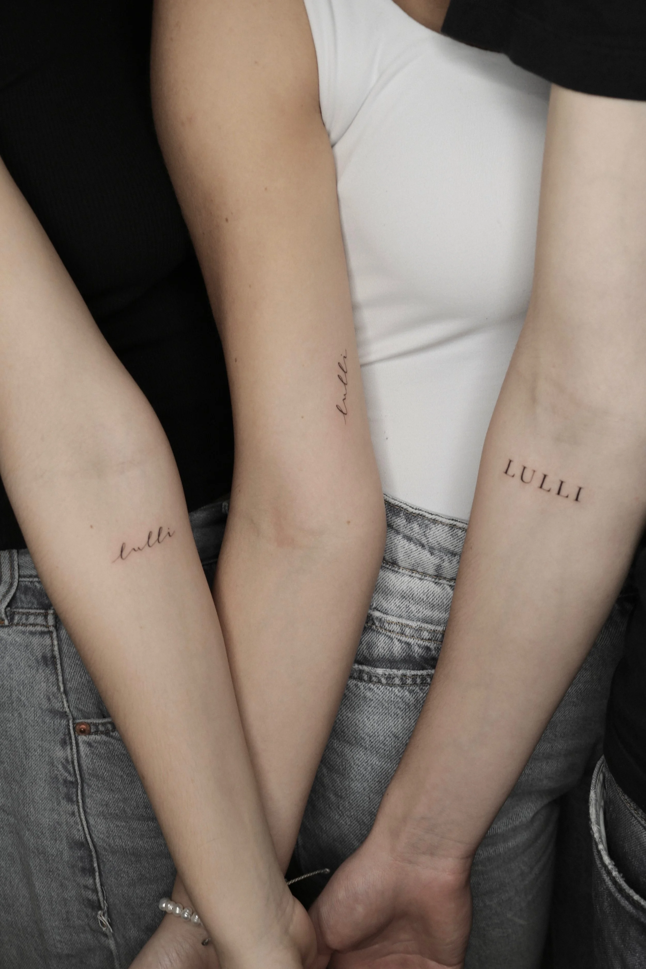 Nahaufnahme von drei Unterarmen mit minimalistischen Tattoos des Wortes "Lulli" in verschiedenen Schriftarten. Die Personen tragen Jeans und Oberteile in Schwarz und Weiß.