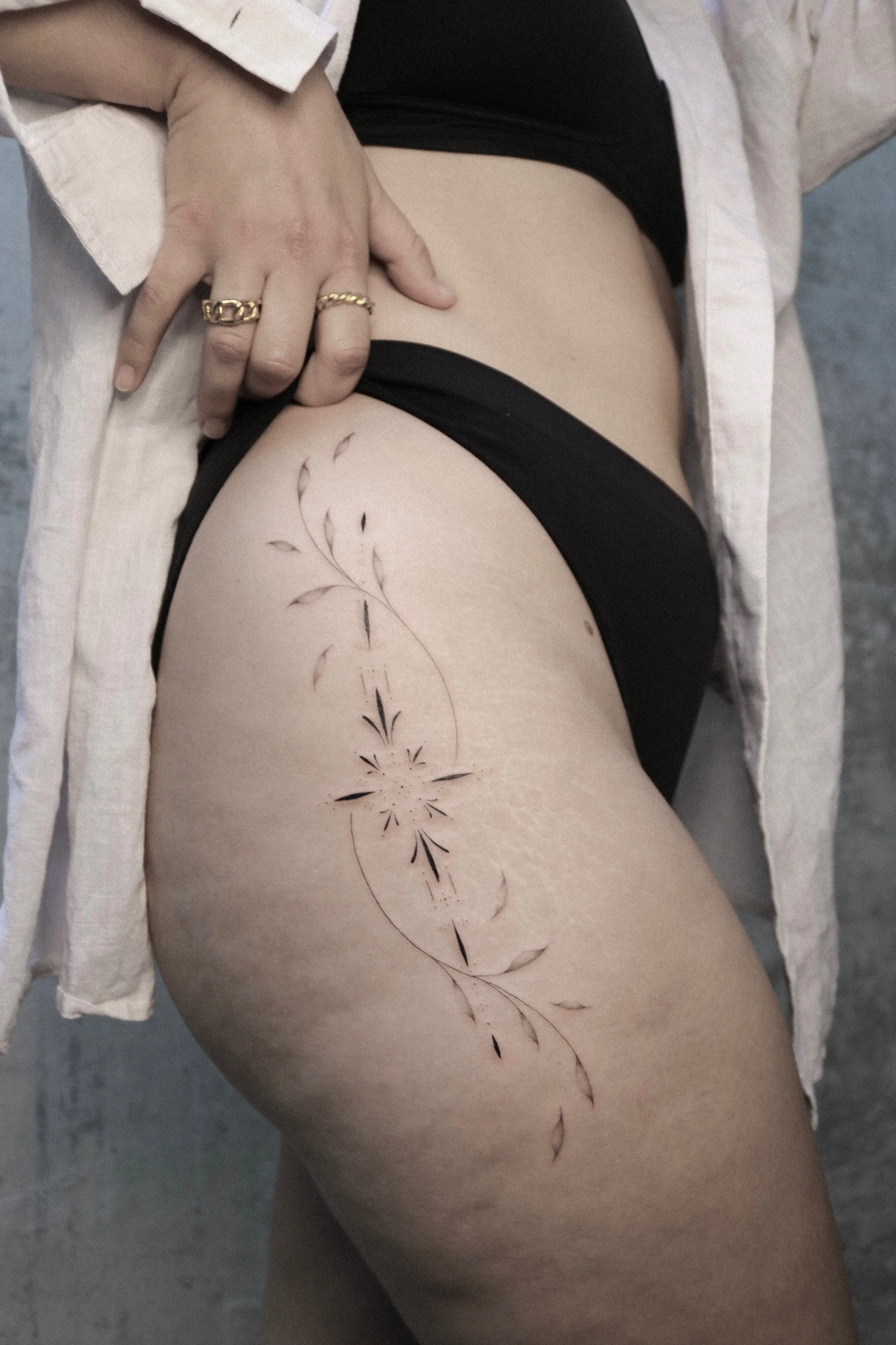 Frau mit floralem Tattoo auf dem Oberschenkel, trägt schwarze Unterwäsche und weißen offenen Mantel, mit goldenen Ringen an der Hand.
