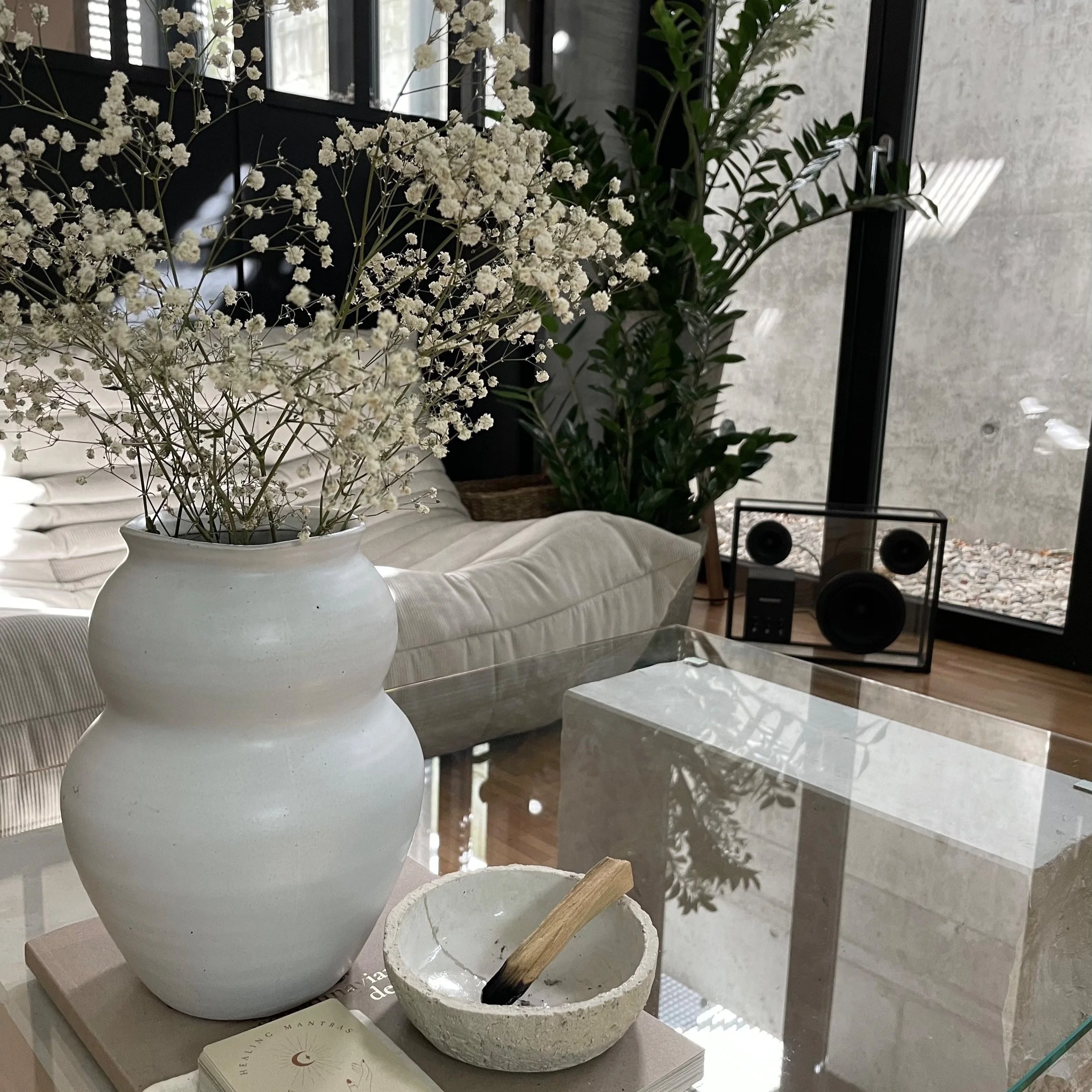 Moderne Wohnzimmerdekoration mit weißer Vase, Trockenblumen und abstraktem Kunstobjekt auf einem Glastisch, weiche Couch im Hintergrund, große Zimmerpflanze und Fenster mit Blick nach draußen.