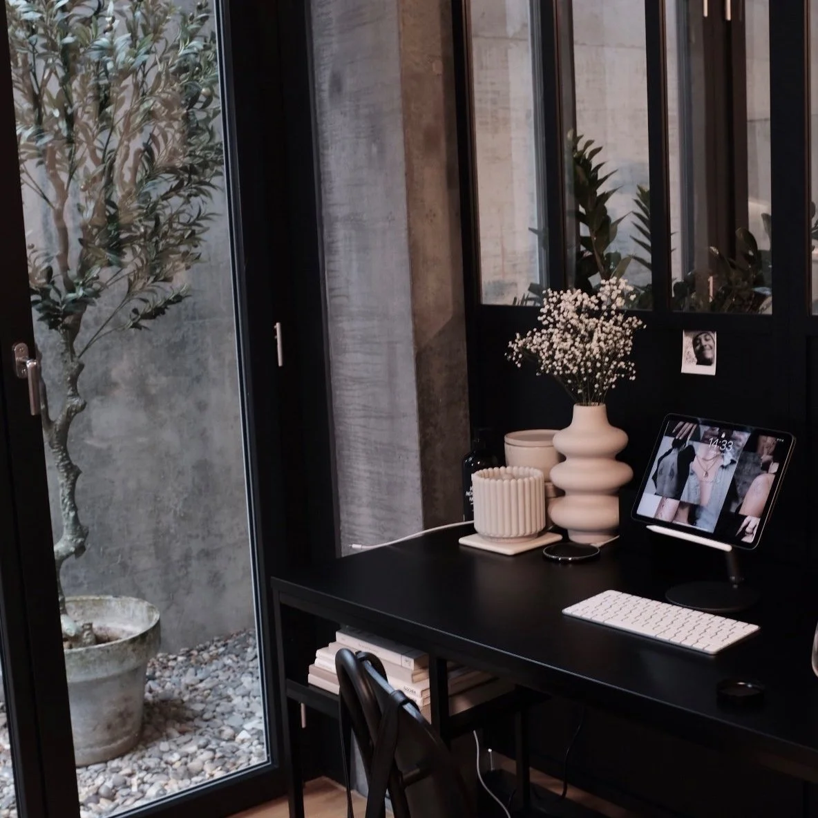Modern eingerichtetes Heimbüro mit schwarzem Schreibtisch, darauf eine Vase mit weißen Blumen, ein Tablet, eine weiße Tastatur und Dekorationen. Durch das Fenster sieht man einen Baum in einem Topf.