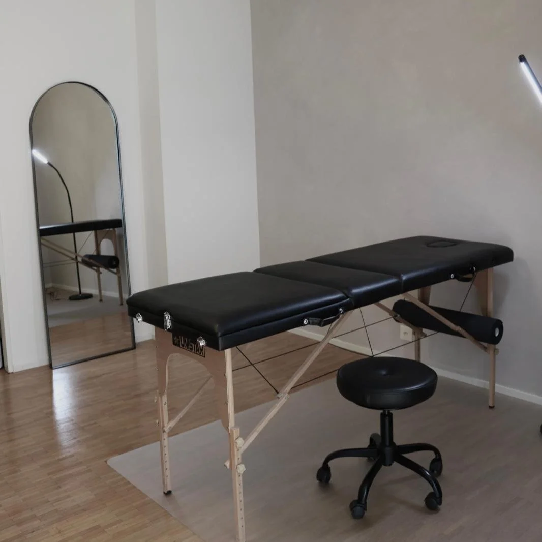 Massagetisch in einem Raum mit Spiegel und schwarzem Hocker vor beiger Wand.