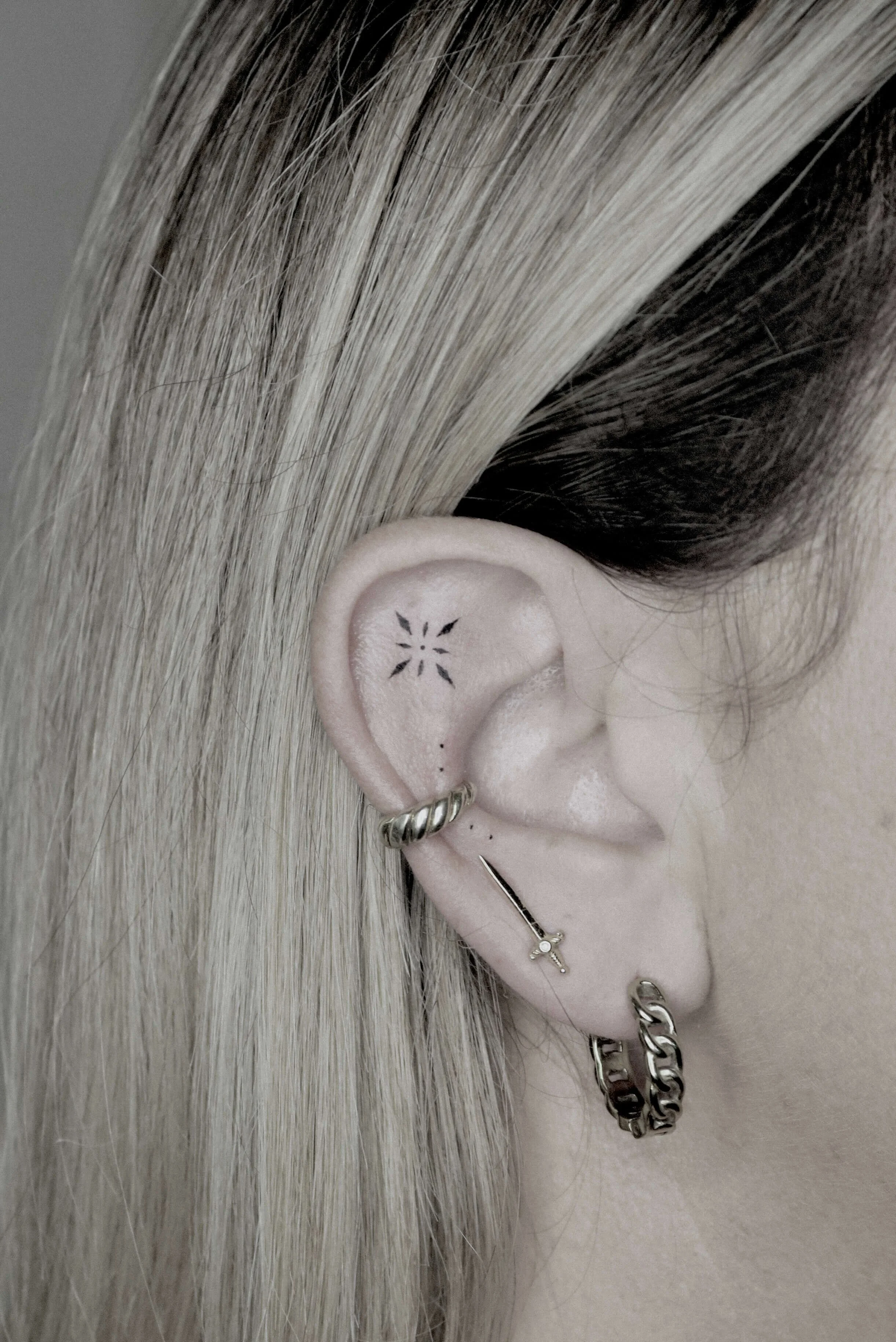 Nahaufnahme eines Ohrs mit mehreren Piercings und kleinen Tätowierungen. Das Ohr hat ein Spiralförmiges, ein Schwert- und ein Kettchen-Ohrpiercing. Das Haar ist blond.