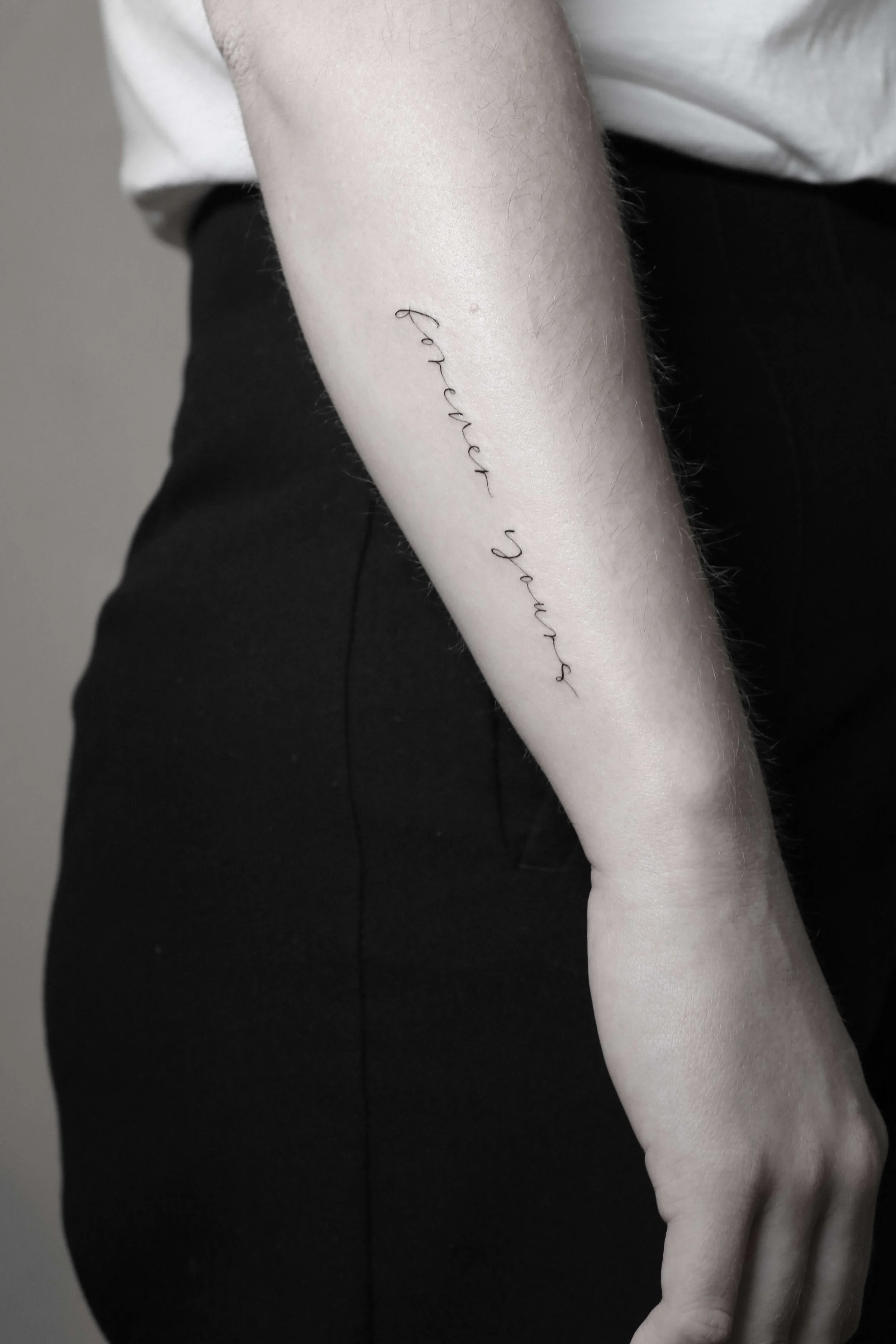 Nahaufnahme eines Unterarms mit einem Tattoo-Schriftzug "forever yours" in schwarzer Tinte.