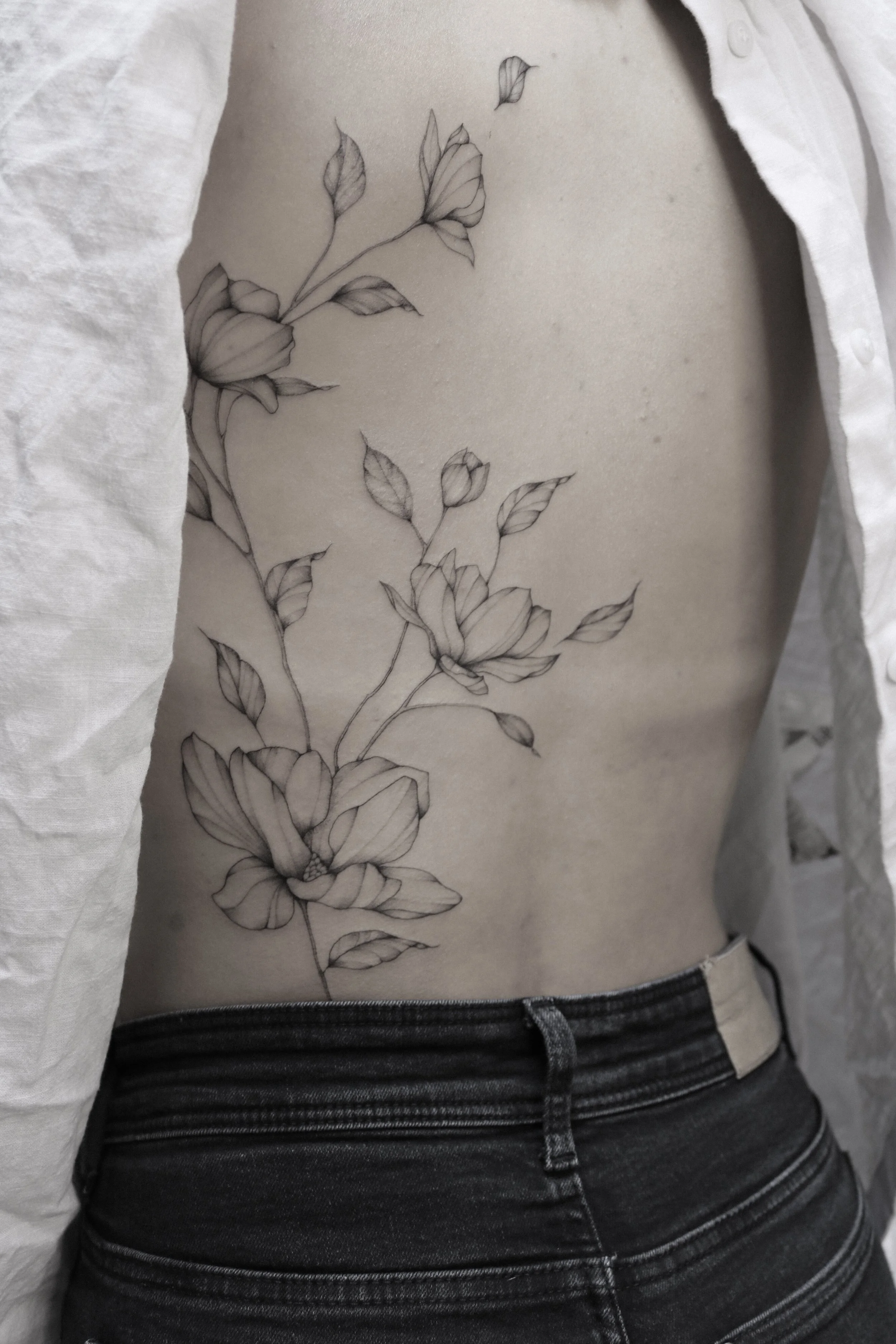 Großes Blumen-Tattoo auf dem Rücken einer Person in Jeans und offenem weißen Hemd.