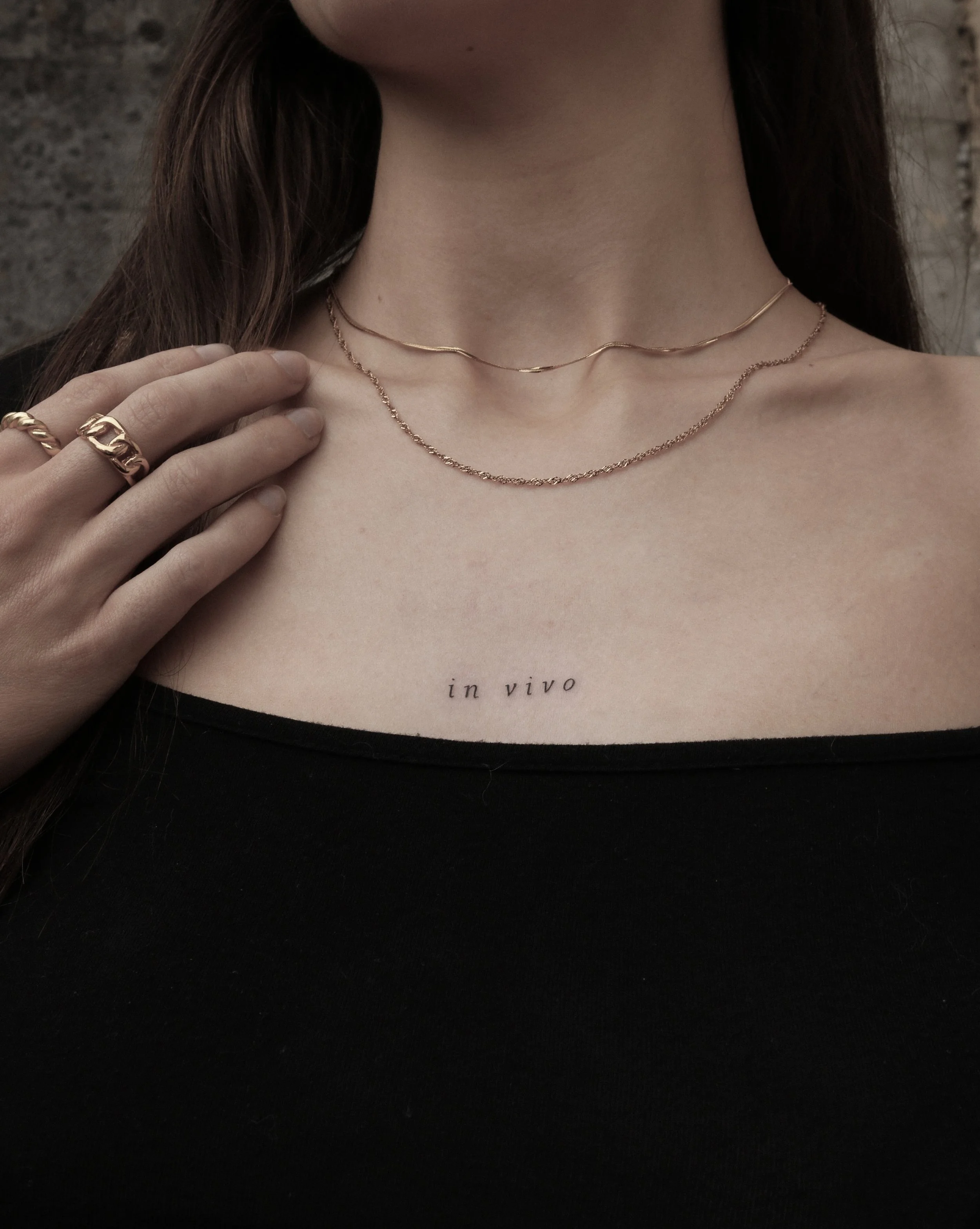 Nahaufnahme eines Halses mit zwei goldenen Halsketten, einem „in vivo"-Tattoo und einer Hand mit zwei goldenen Ringen auf schwarzem Oberteil.