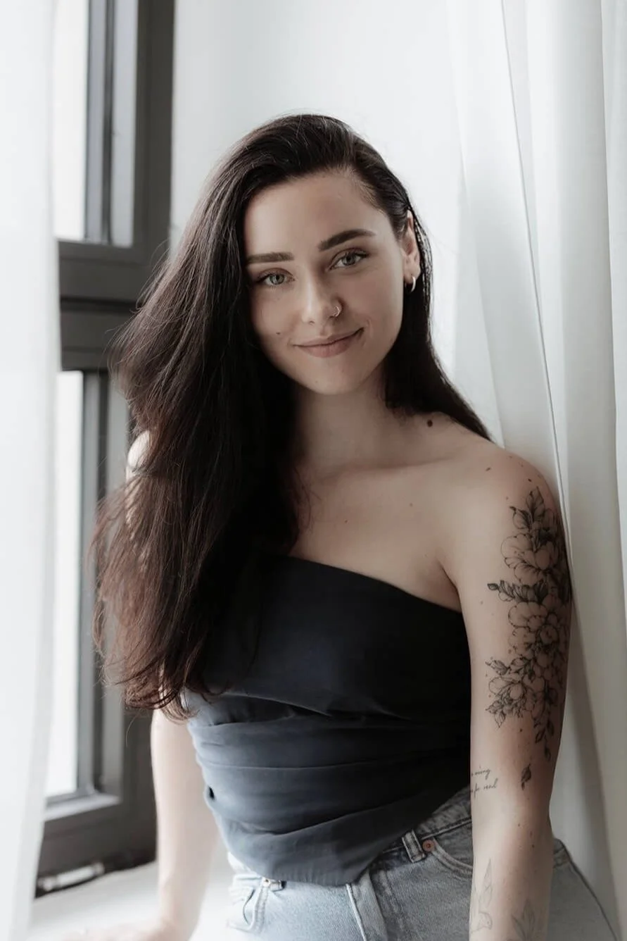 Frau mit langen Haaren, Tattoo am Arm, steht lächelnd am Fenster, trägt ein schwarzes Oberteil und Jeans.