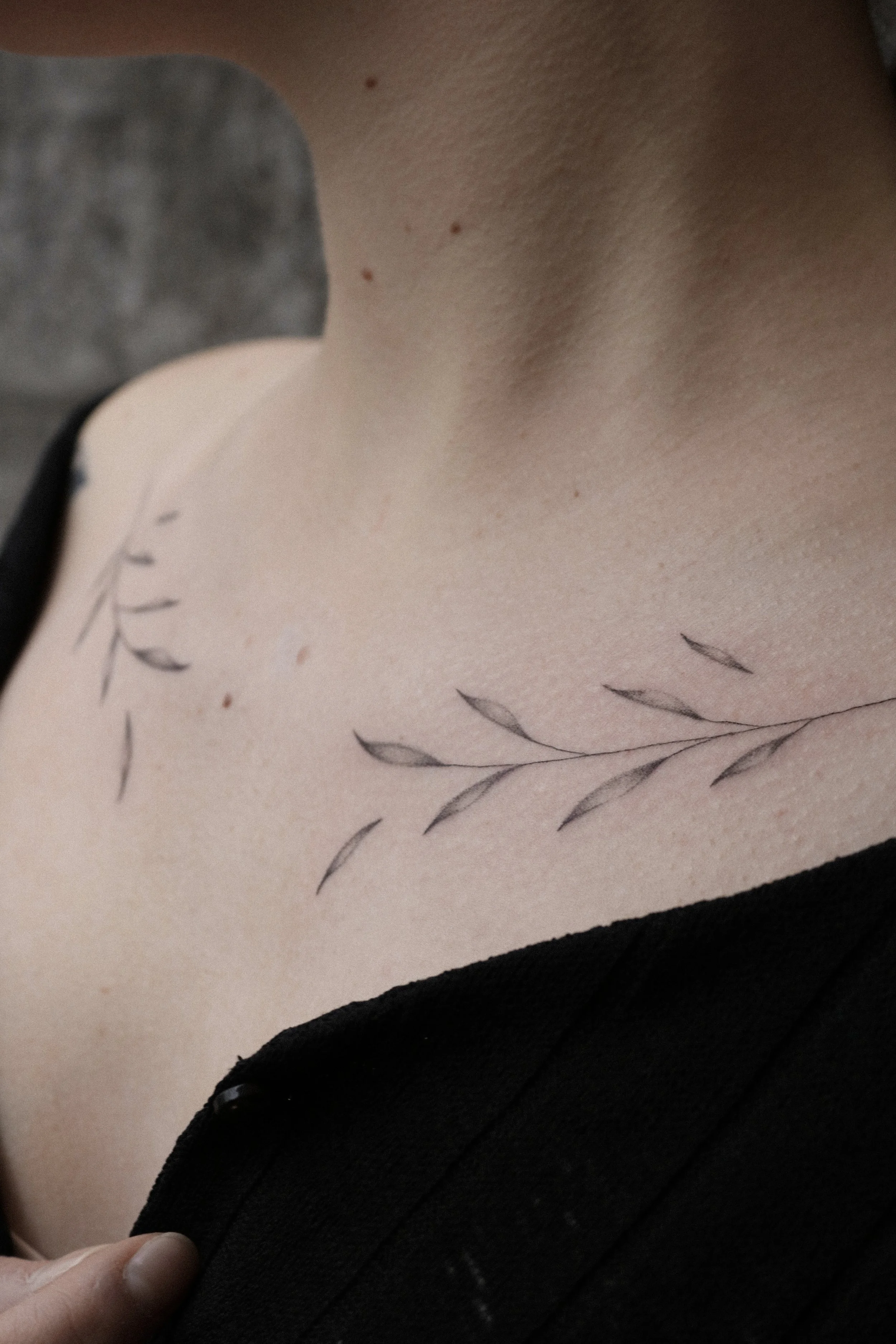 Feines Zweig-Tattoo auf der Schulter einer Person, in schwarz-weißem Design, mit heller Haut und einem schwarzen Kleid.