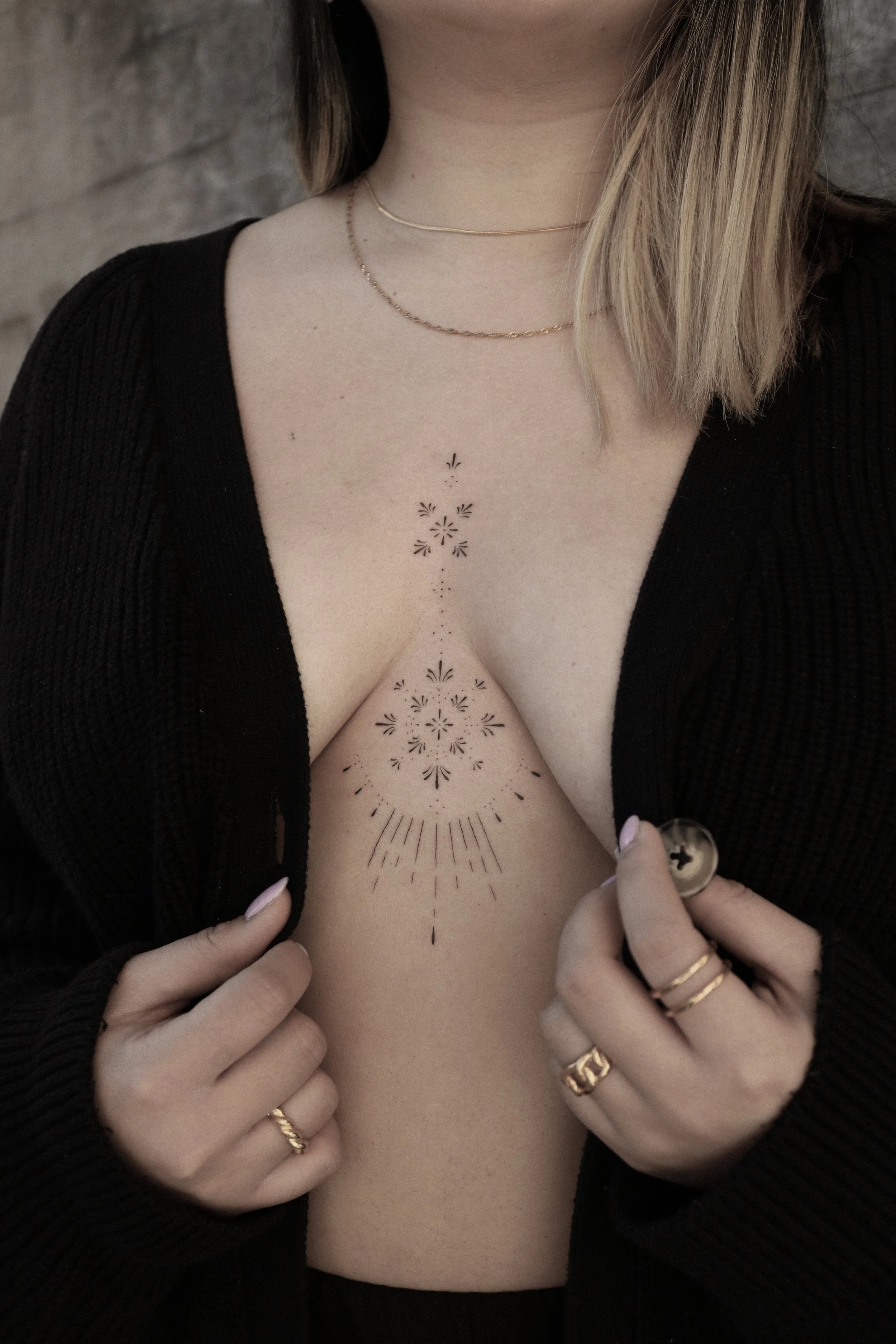 Frau mit Dekolleté-Tattoo und Goldschmuck, trägt schwarzen Cardigan, Tattoo in geometrischen Mustern.