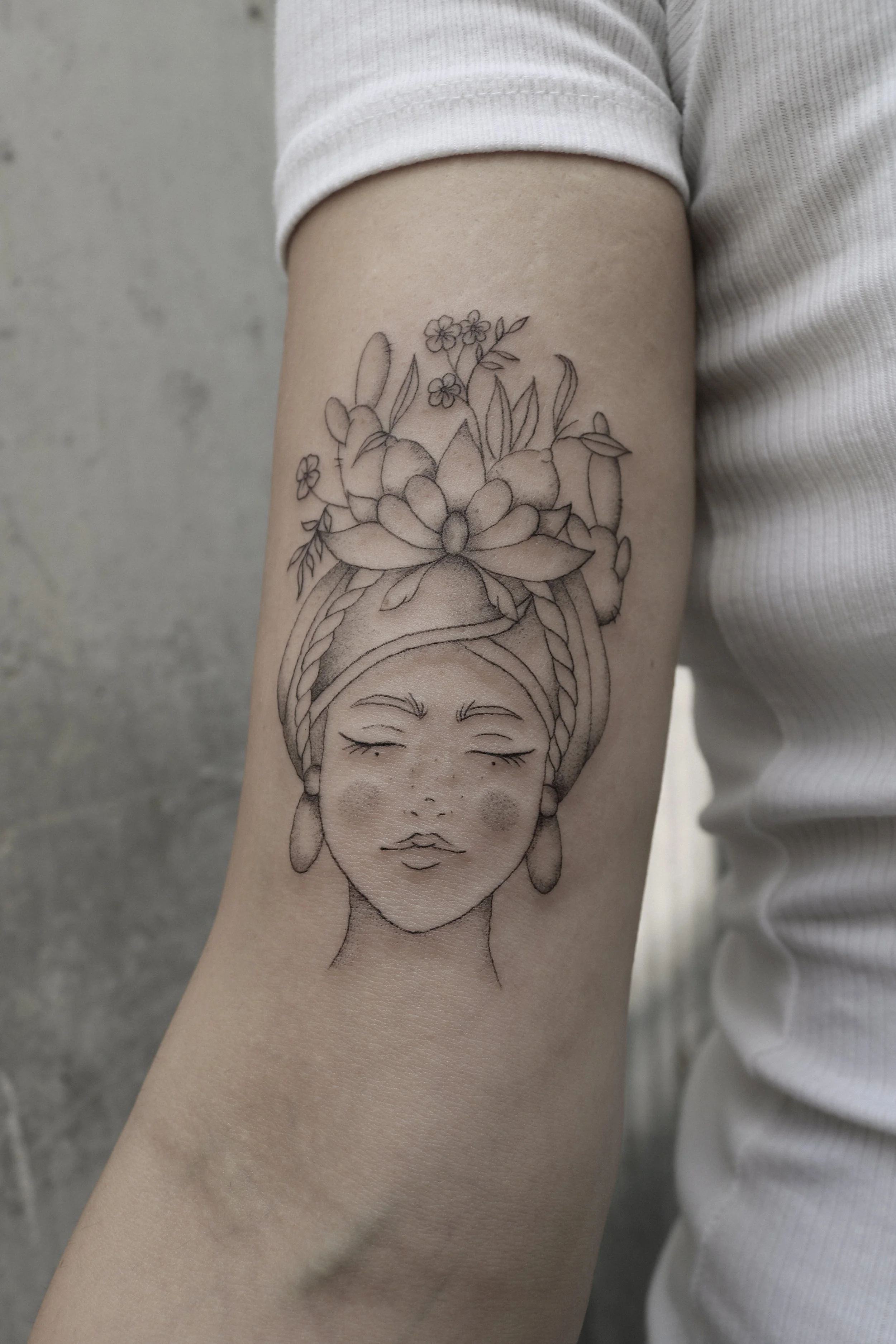 Tätowierung auf einem Arm mit dem Gesicht einer Frau, geschlossenen Augen und Blumen im Haar. Die Frau trägt ein Kopftuch und Ohrringe. Das Motiv ist in Schwarz-Weiß gehalten.