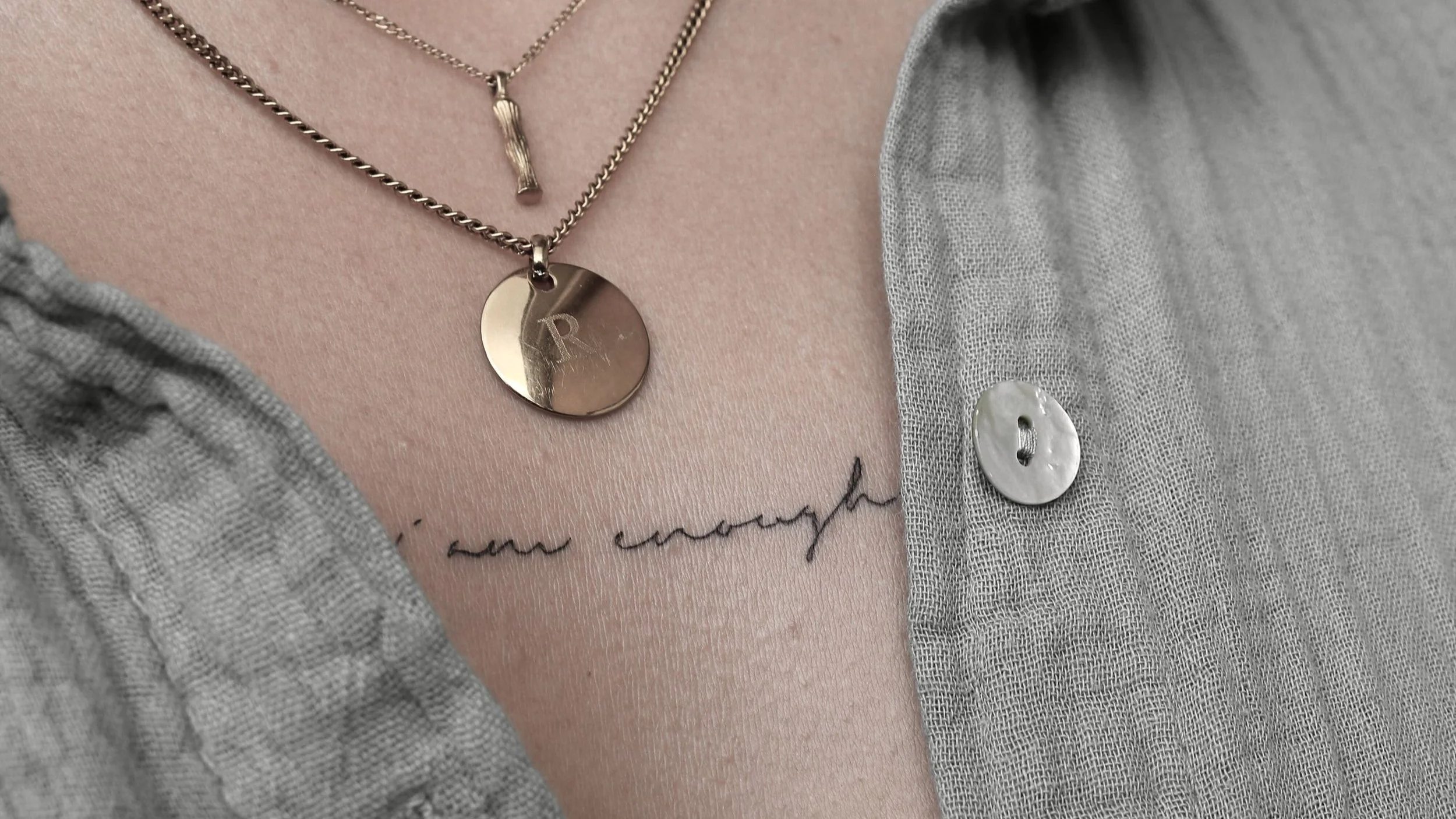 Nahaufnahme eines Tattoos "I am enough" auf der Haut mit zwei goldenen Halsketten und einem grauen Hemd mit Knopf. Stilvoll und minimalistisch gestaltet.