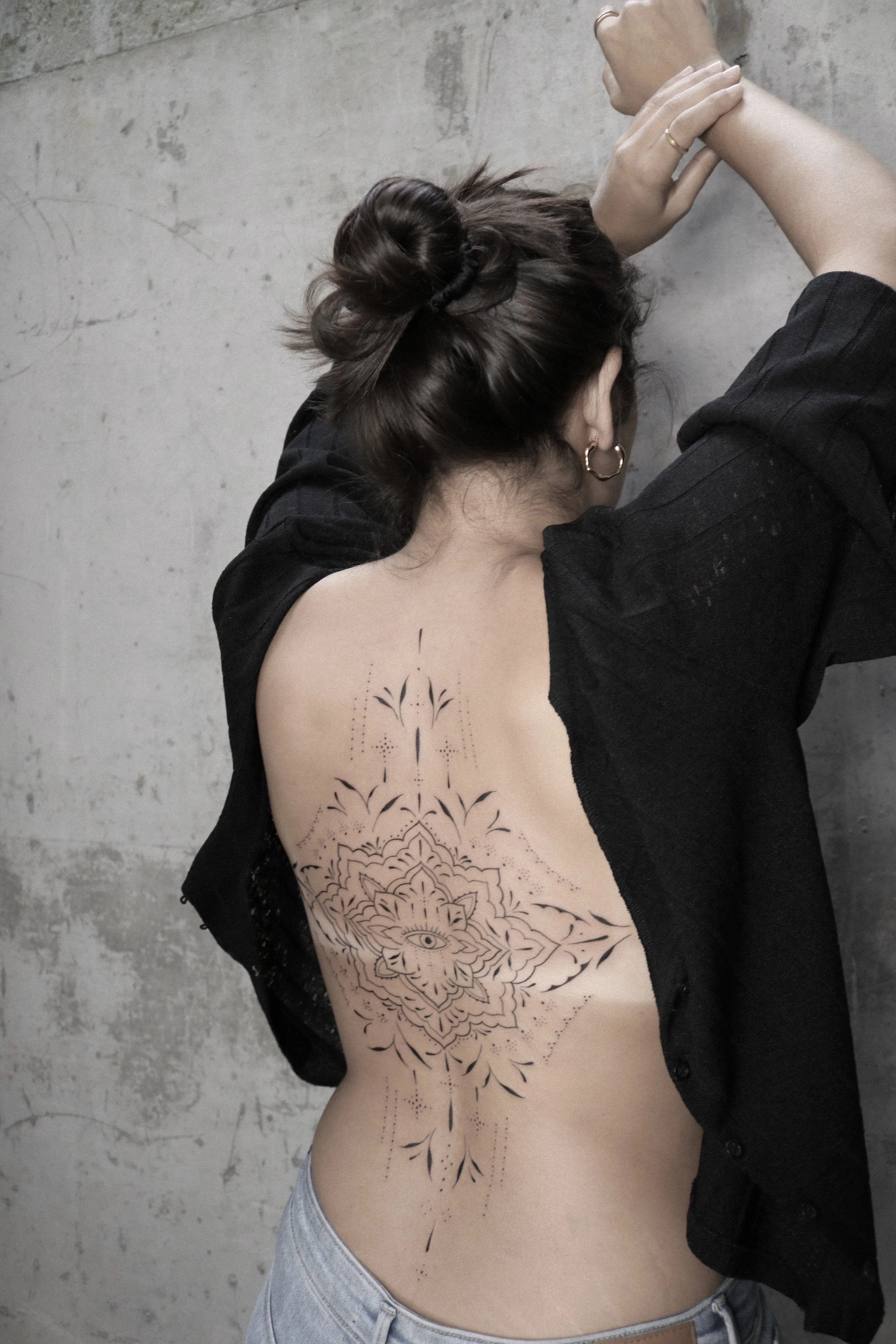 Frau mit Rückentattoo, geometrisches und florales Design, trägt schwarze Kleidung und Jeans, Haar hochgesteckt, vor Betonwand.