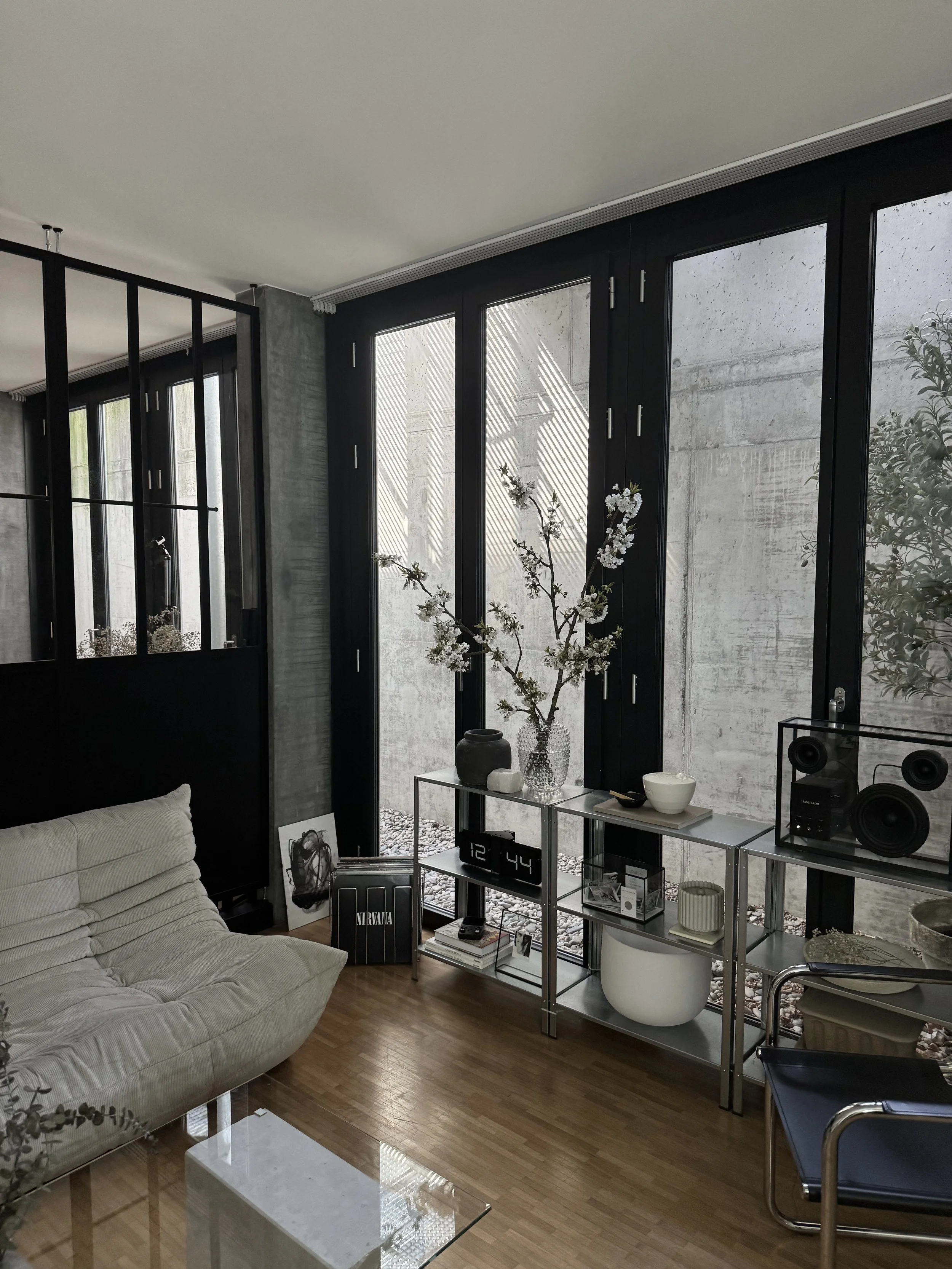 Modernes Wohnzimmer mit bequemer, hellgrauer Couch, Glas-Beistelltisch und Regalen mit Dekorationen. Große Fenster lassen Tageslicht herein und ein minimalistischer Einrichtungsstil ist erkennbar.