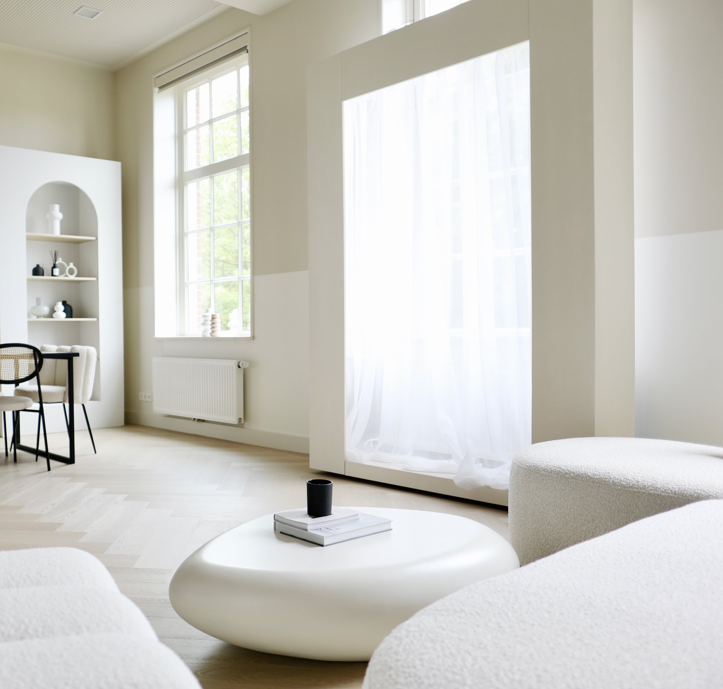 Minimalistisch ingerichte woonkamer met witte meubels, een lage salontafel met een boek en een kaars, en een grote raam met lichte gordijnen. Er is ook een open kast met decoratieve objecten.