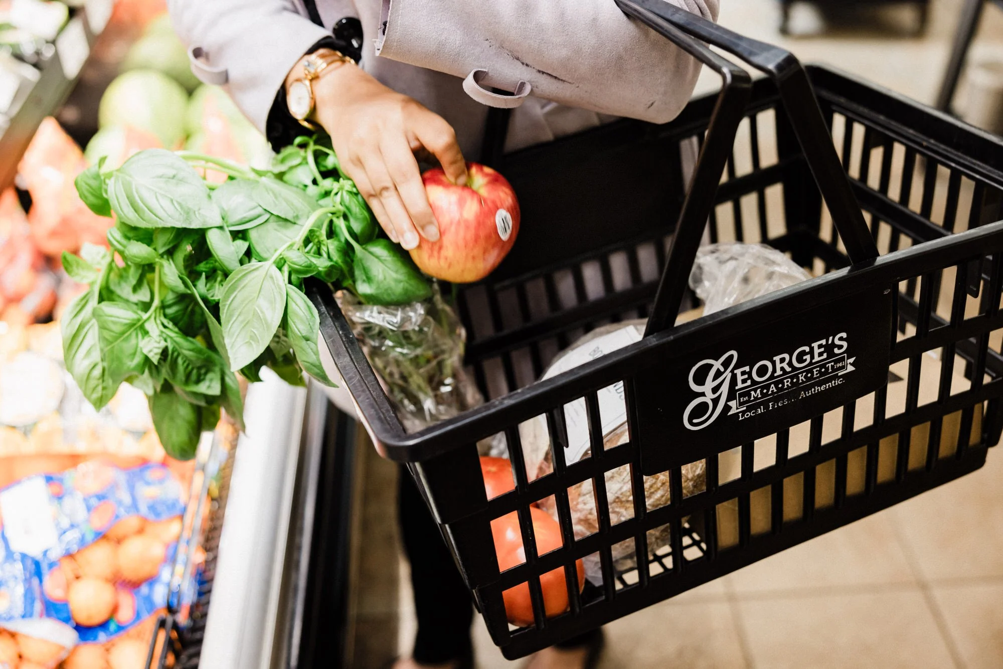 A person shopping at a grocery store placing an apple and fresh basil into a shopping basket labeled George's Market.