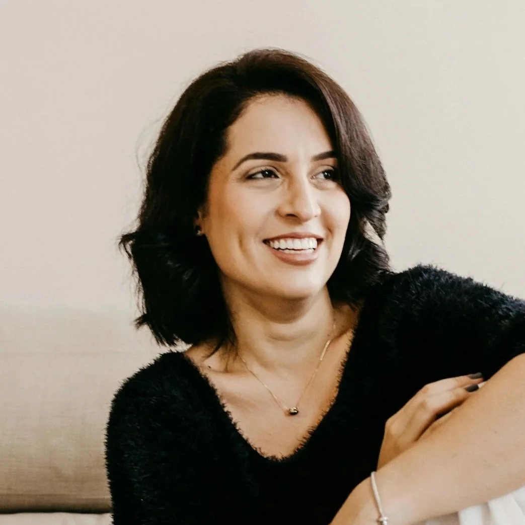 A woman with shoulder-length dark hair smiling, wearing a black fuzzy sweater, a delicate gold necklace, and a bracelet, sitting on a light-colored couch against a plain beige wall.
