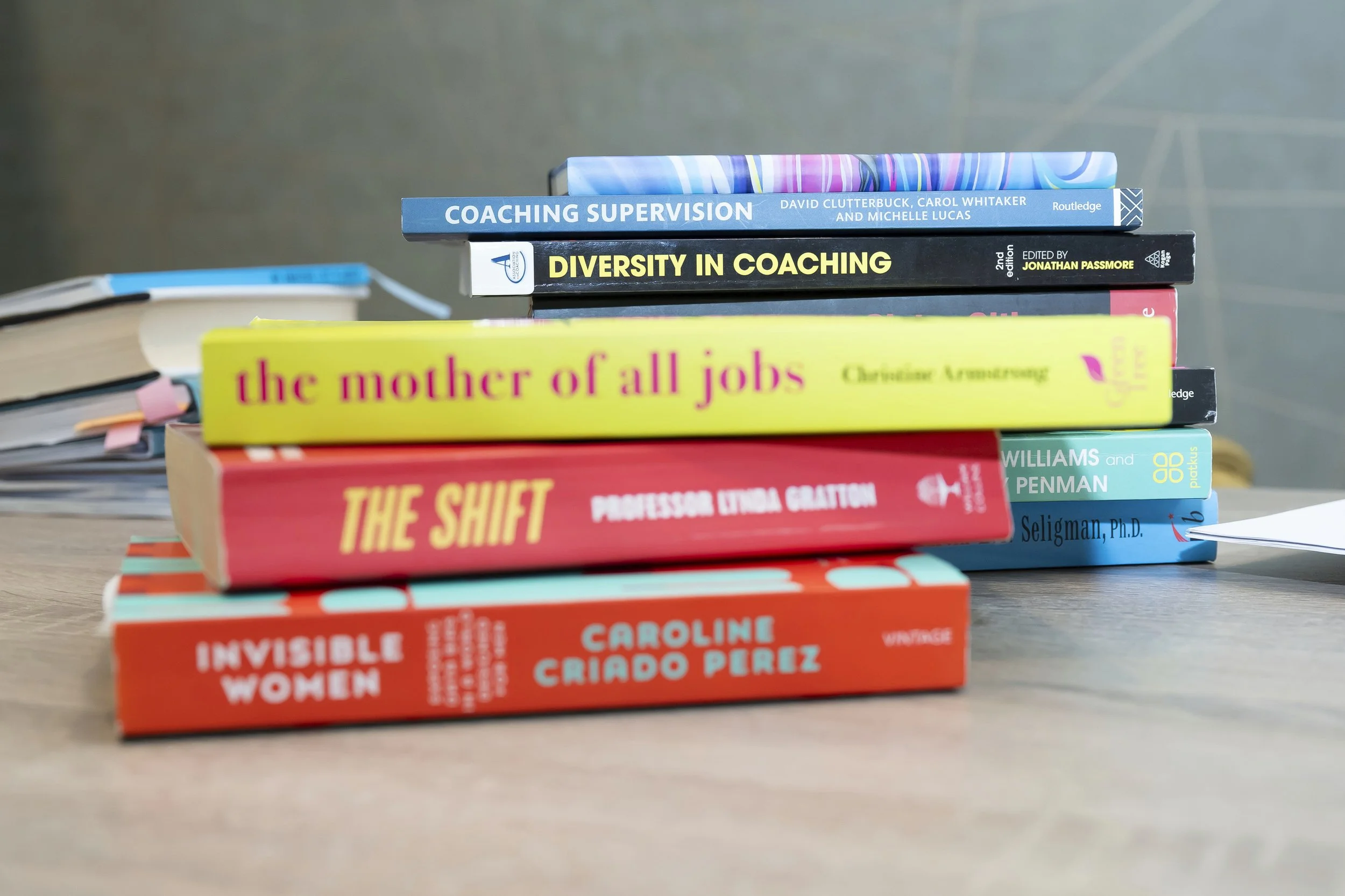 A stack of books on a wooden table, with additional books and papers in the background. The visible titles include 'Coaching Supervision,' 'Diversity in Coaching,' 'The Mother of All Jobs,' 'The Shift,' and 'Invisible Women.'