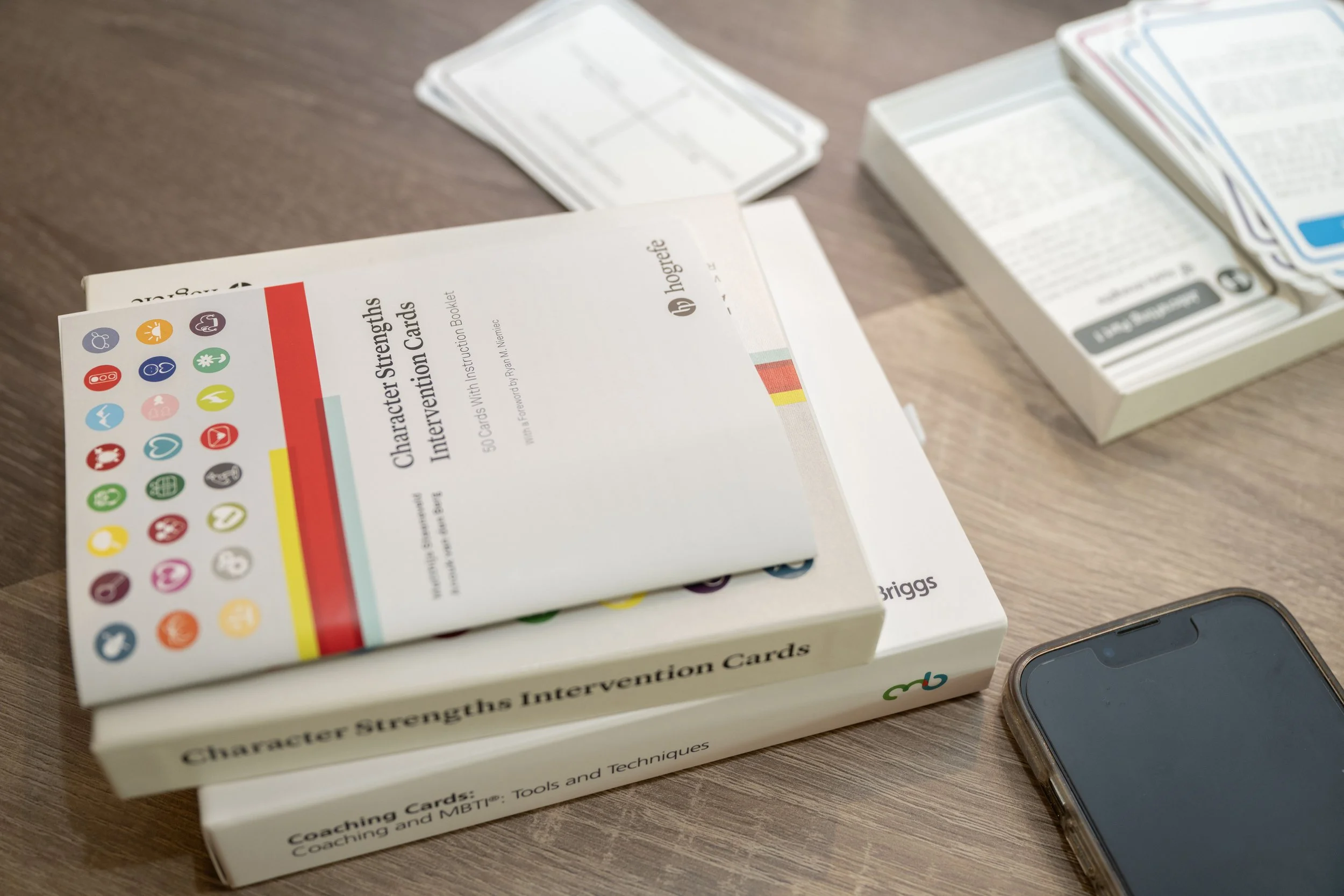 Books titled "Character Strengths Intervention Cards" and "Coaching Cards: Tools and Techniques" on a wooden table, with a box of strengths and MBTI cards.