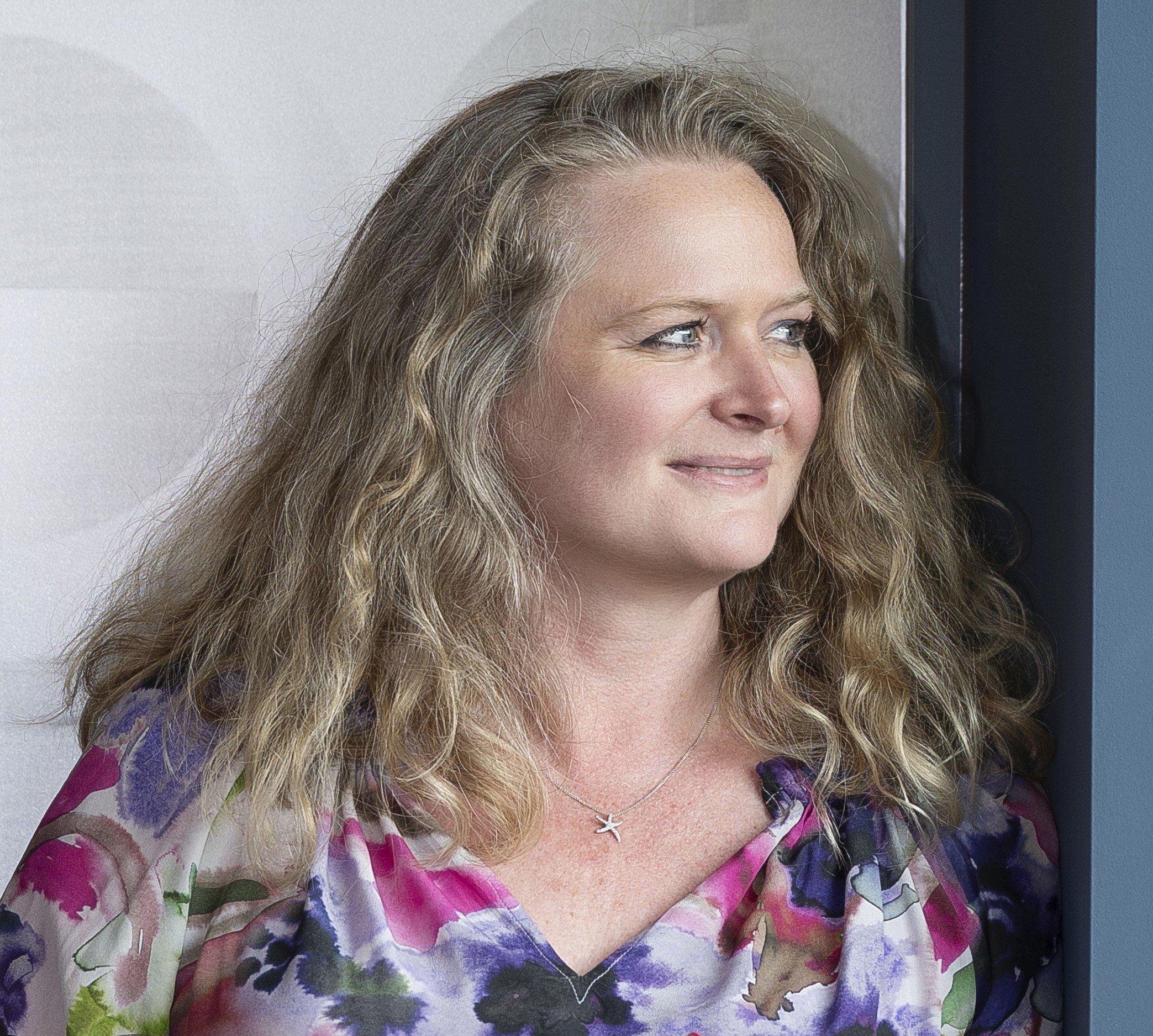 A coach with blond, wavy hair looking to her right, standing near a wall with a purple and blue color scheme, wearing a colorful floral blouse and a starfish necklace.