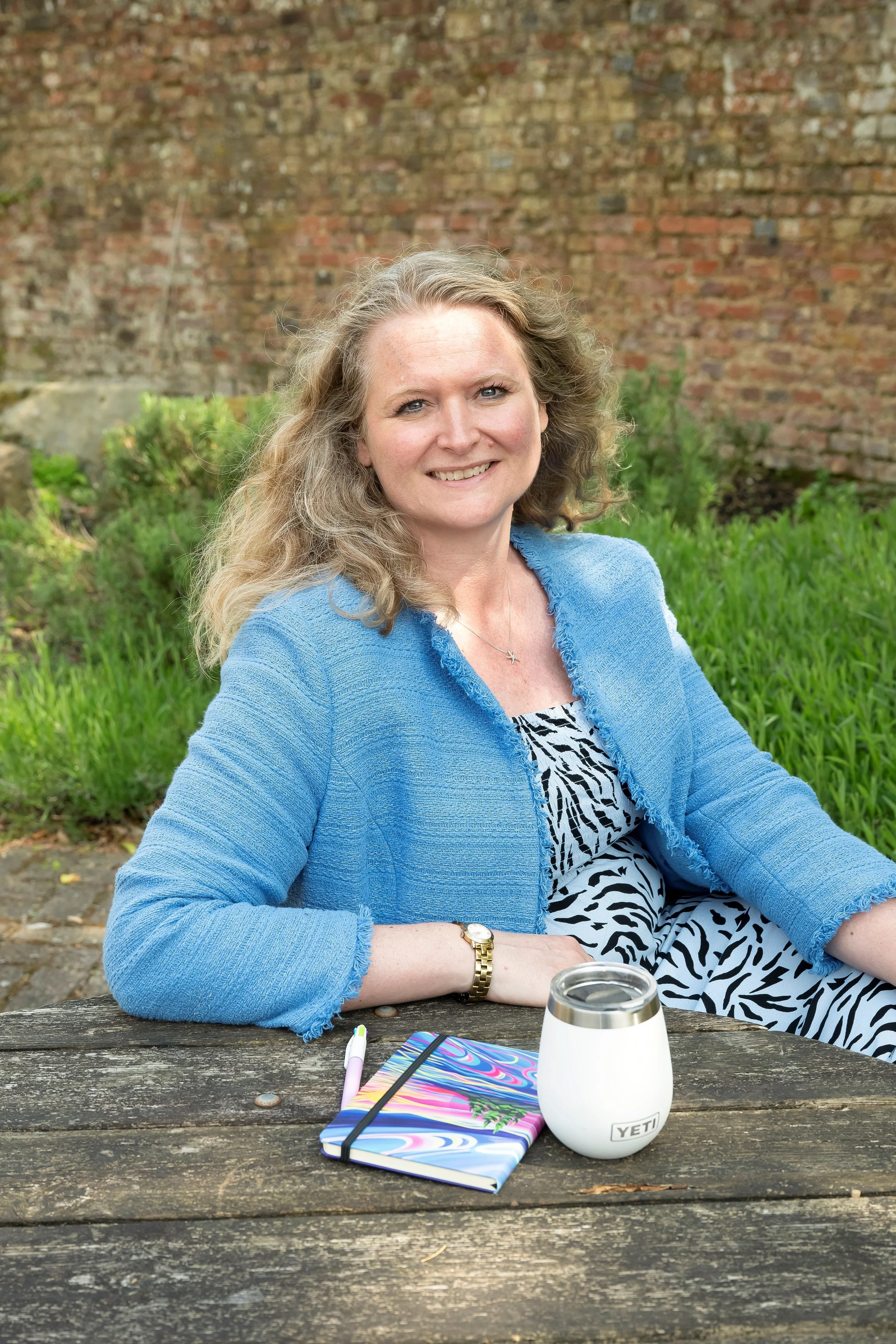 A coach with long curly blonde hair and blue eyes smiling at the camera, wearing a blue jacket with a brick wall and greenery in the background.
