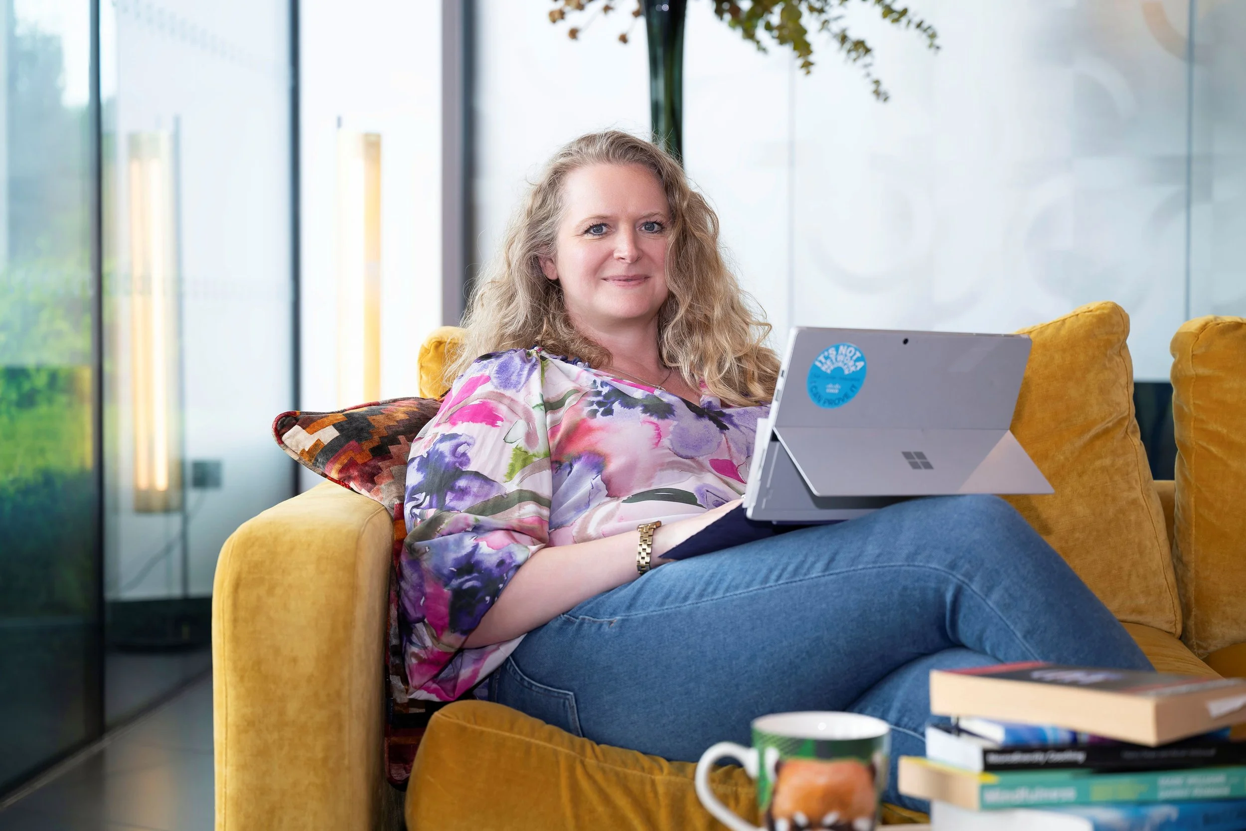 A woman with curly blonde hair sitting on a mustard yellow couch, working on a laptop, wearing a colorful floral blouse and blue jeans, in a modern indoor setting with large windows and a whiteboard in the background.