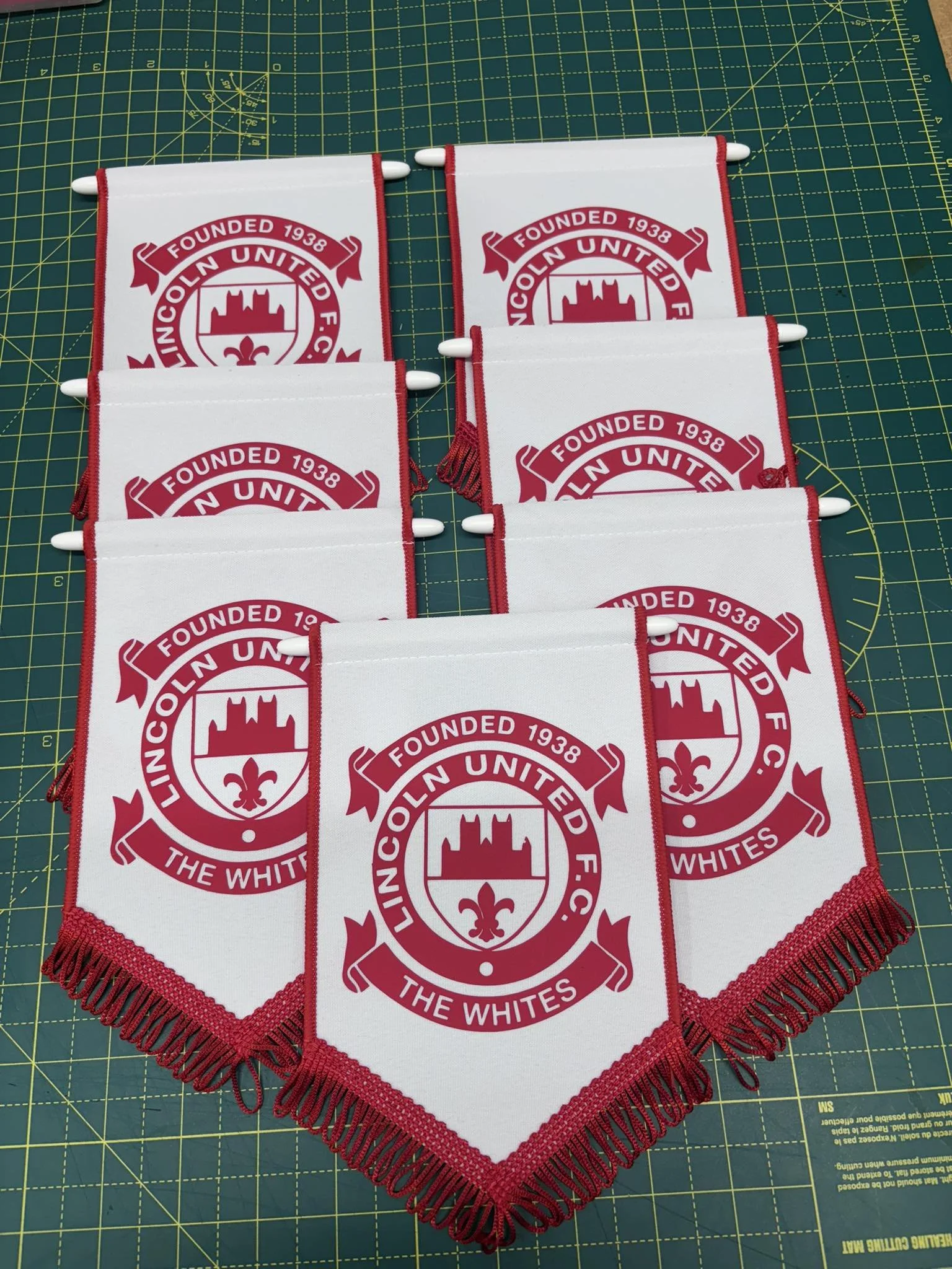 A set of six small flags with Lincoln United F.C. emblem, laid out on a green cutting mat; the flags are white with red borders, fringe, and feature a circular emblem with a castle, a fleur-de-lis, and the text "Founded 1938 Lincoln United F.C. The W