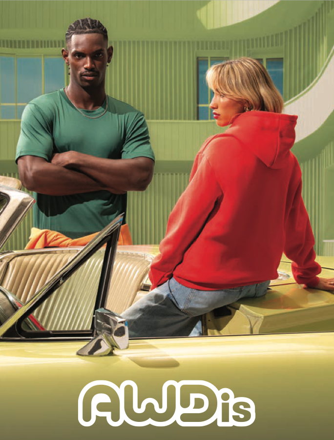 Two people in a brightly colored room, one man with dark skin and braided hair standing with crossed arms wearing a teal shirt, and one woman with light skin and blonde hair sitting on the hood of a yellow car wearing a red hoodie. The scene has a retro vibe with green walls and a curved backdrop.