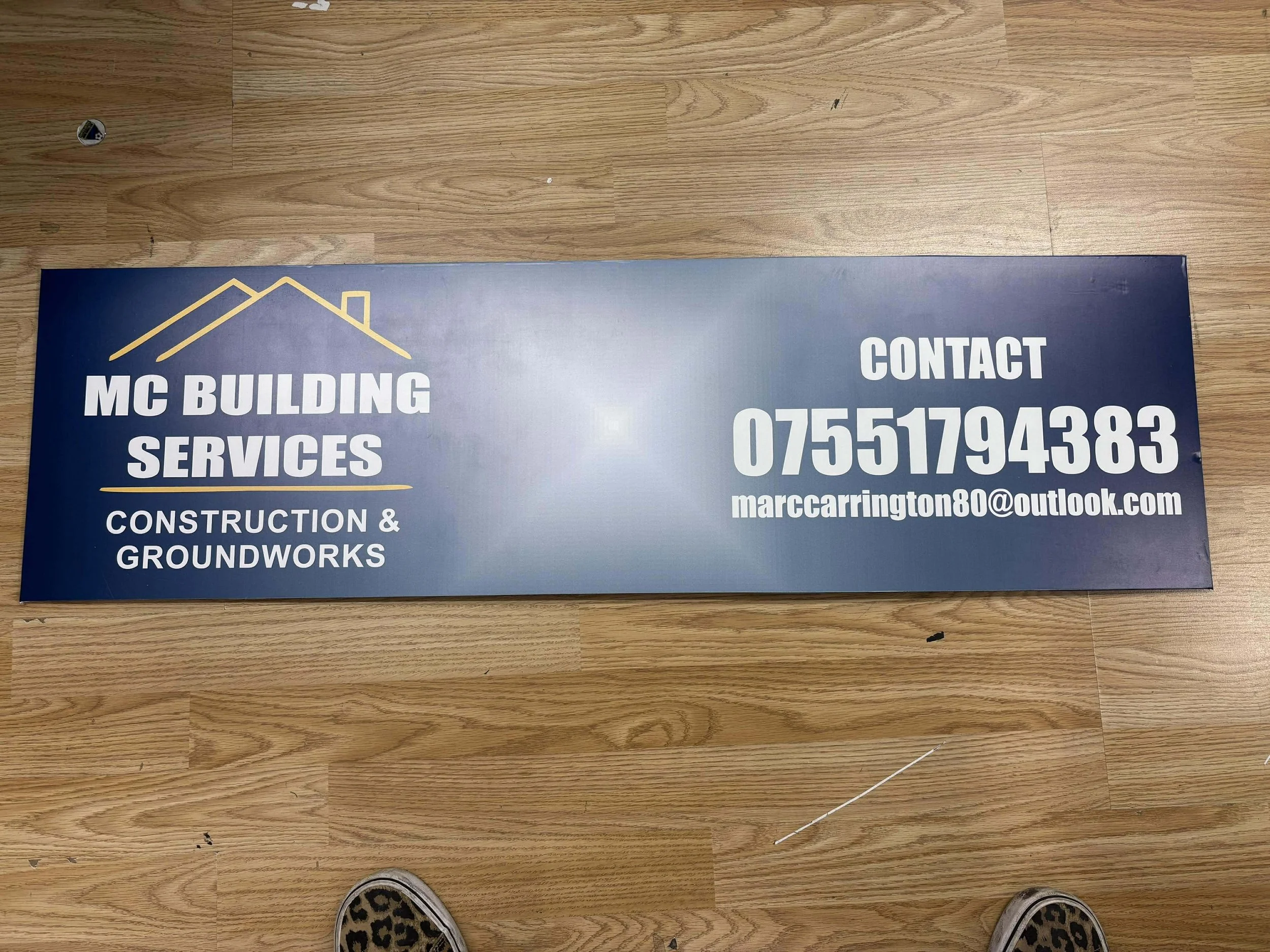 Blue rectangular sign for MC Building Services with a yellow outline of a roof and chimney, including contact information and email on a wood floor.