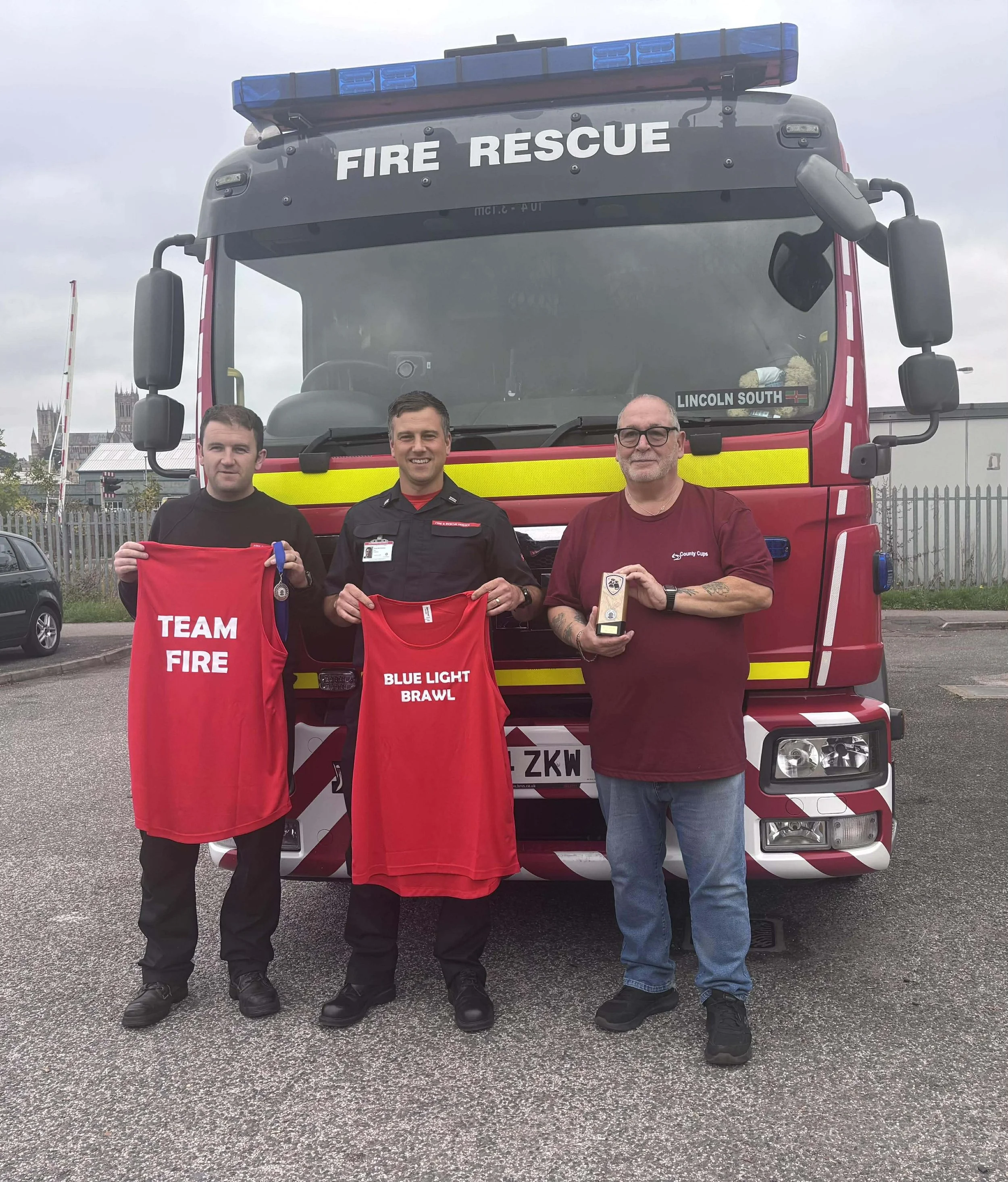 Three men standing in front of a fire rescue truck, holding sports jerseys and a trophy, under a cloudy sky.
