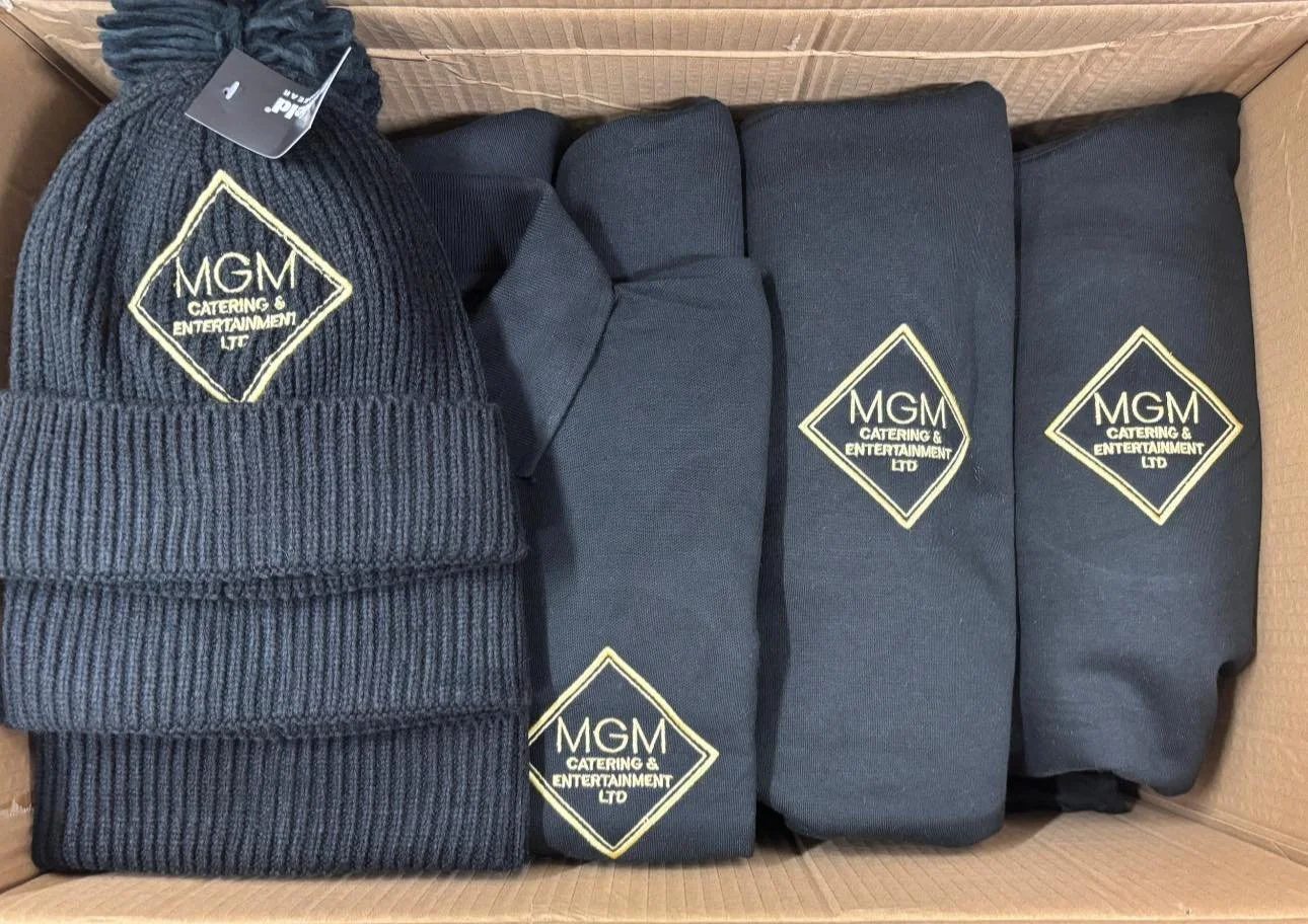 A cardboard box containing several black hooded sweatshirts with a logo that reads "MGM Catering & Entertainment Ltd" and a blue knit beanie hat with the same logo.