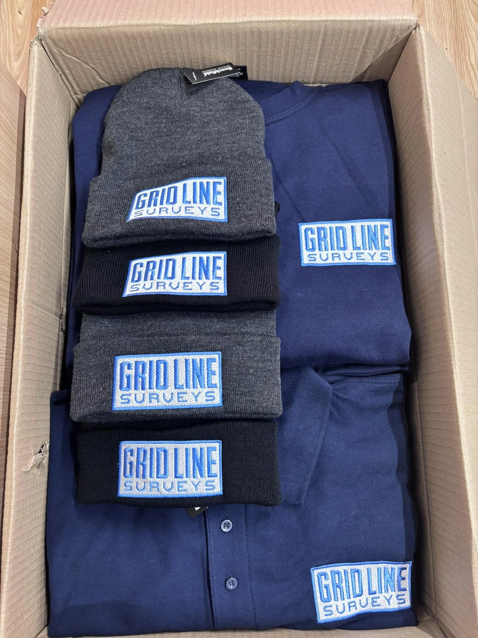 A cardboard box containing identical navy blue caps, folded navy blue shirts, and navy blue patches, all branded with the 'Grid Line Surveys' logo embroidered in blue and white.