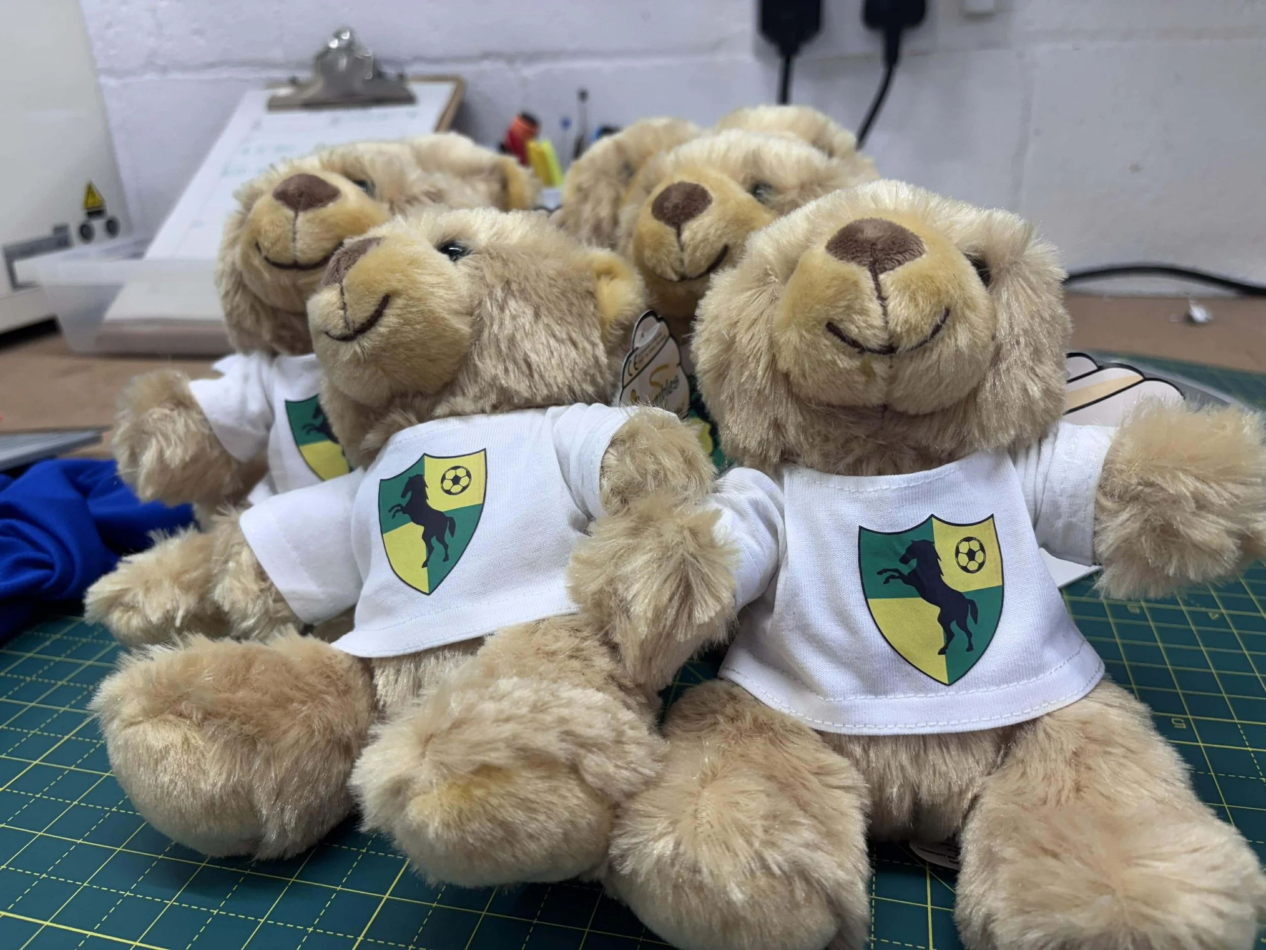 Five plush teddy bears wearing white shirts with a green and yellow shield logo featuring a black horse and a soccer ball on a green cutting mat. They are arranged closely in a workshop or crafting area.
