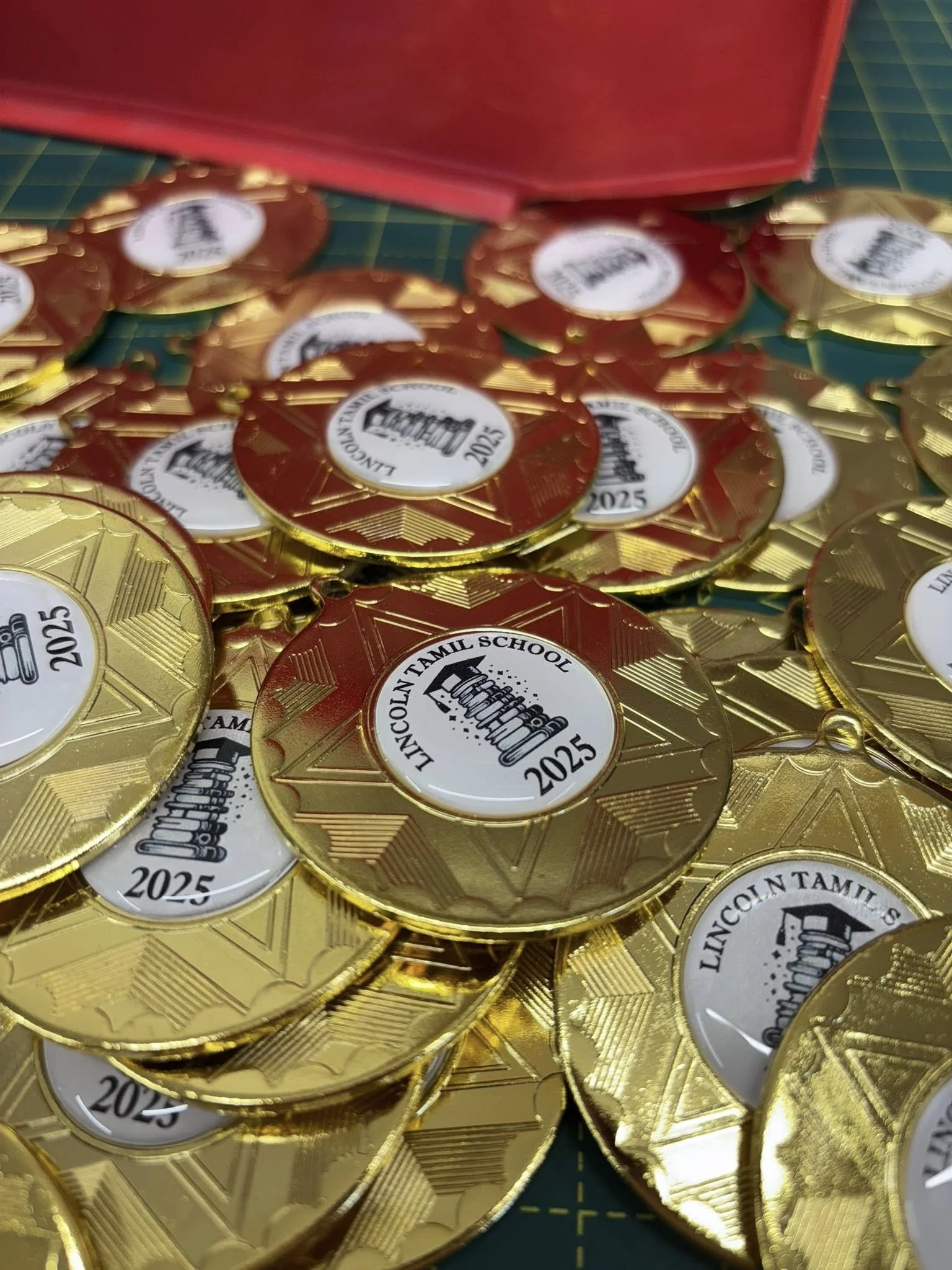 Gold-colored commemorative medals with the Lincoln Tamil School logo and the year 2025 displayed on a white circular background, spread out on a green cutting mat.