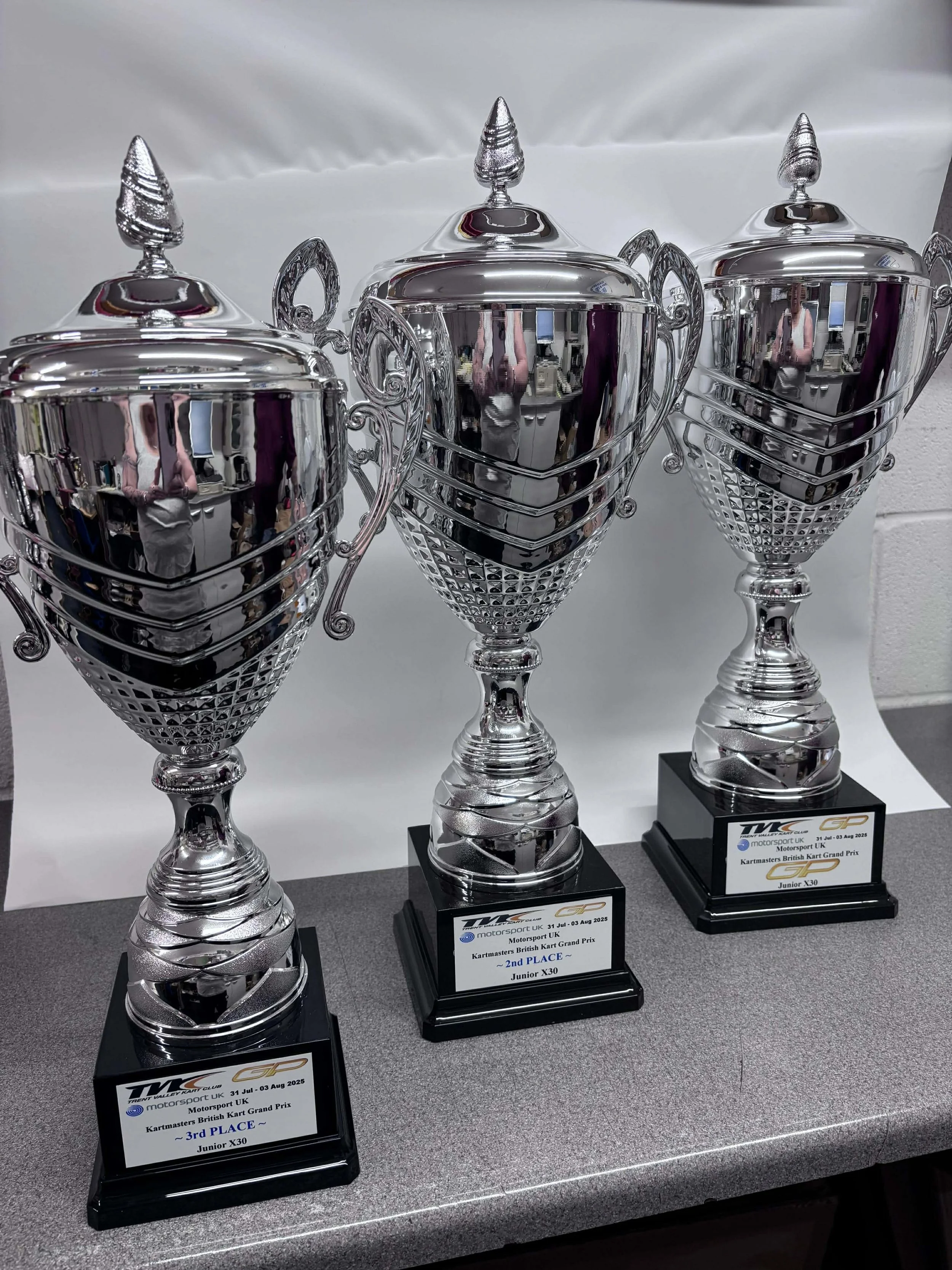 Three shiny silver trophies with black bases and engraved plaques for kart racing, arranged on a gray table indoors.